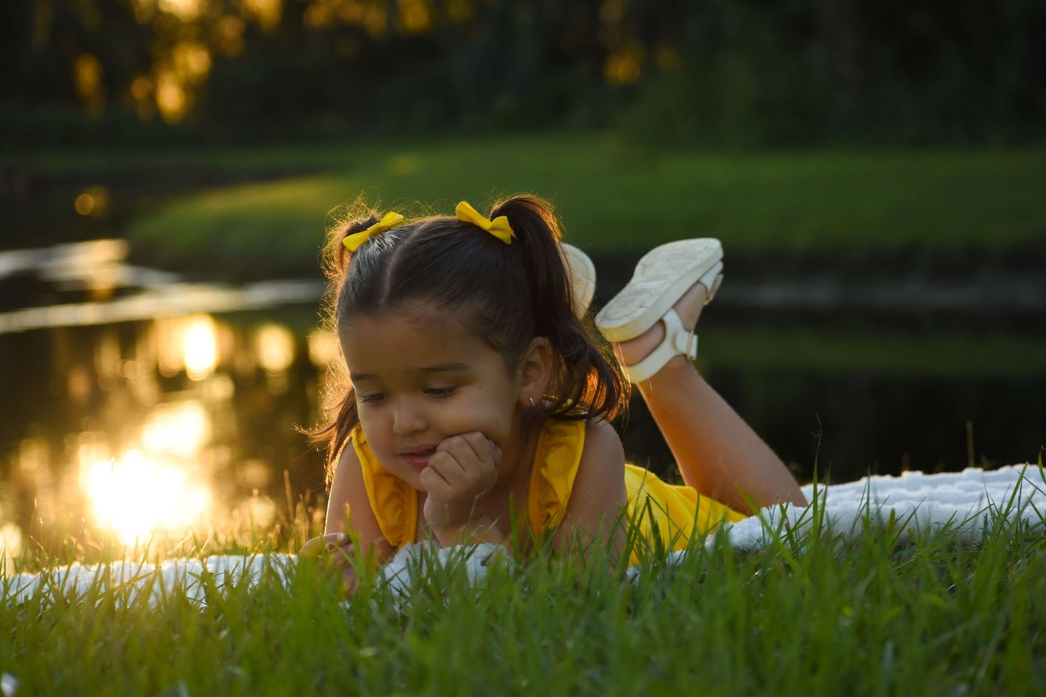 Young girl in yellow dress lying on grass reading a book near a pond at golden sunset.