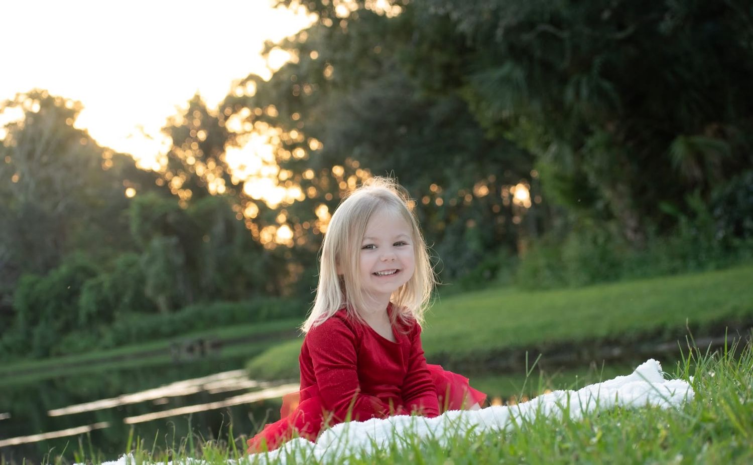 Happy blonde little girl in red dress sitting on green grass with sunset bokeh background.