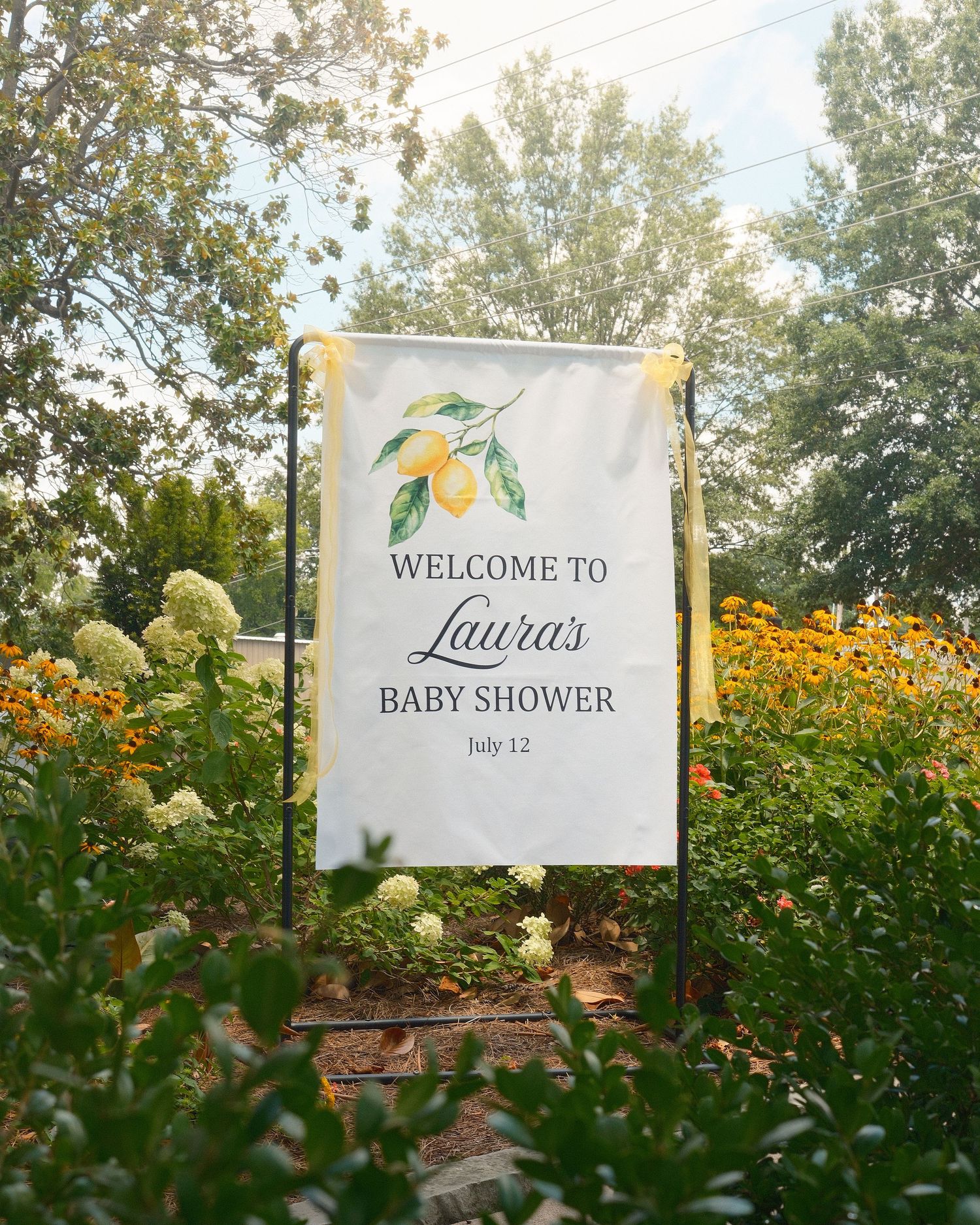 A baby shower banner in the middle of a yellow flowery garden.
