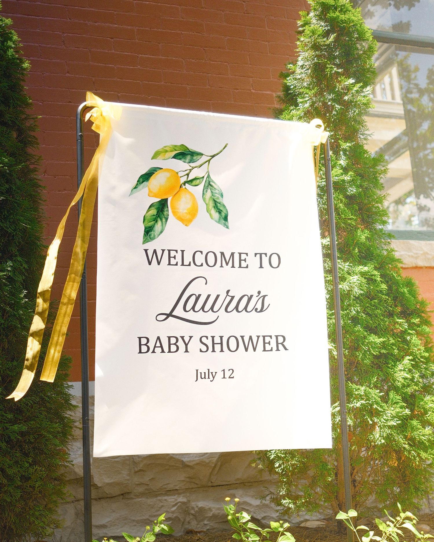 A baby shower banner against a brick wall with greenery surrounding it.
