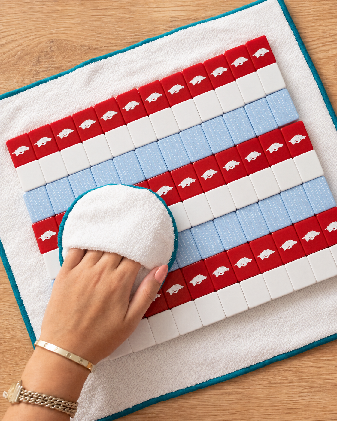 A product photo of mahjong tiles being wiped down by hand on a warm oak tabletop.