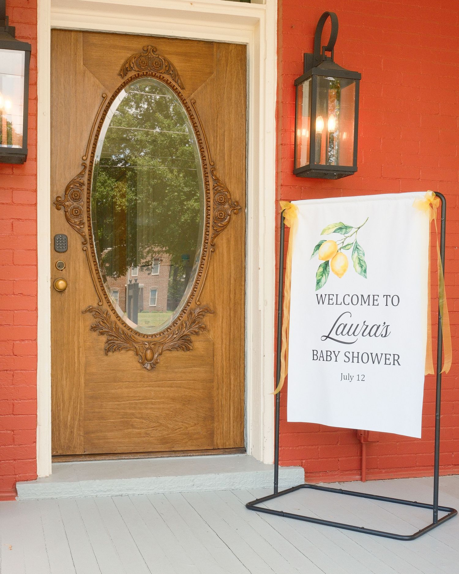 A baby shower banner on the porch of a building against a red brick wall next to a wooden door.