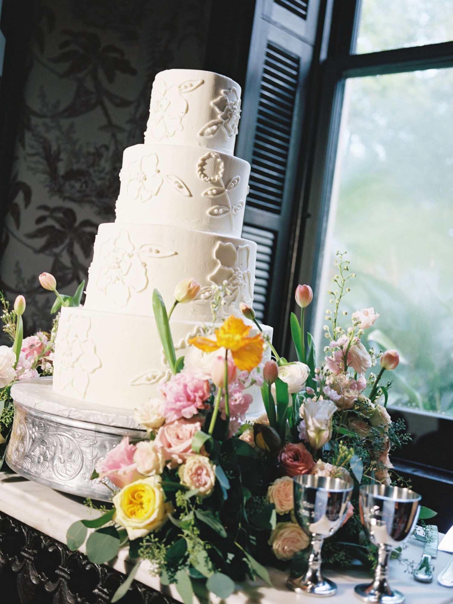Four-tier white wedding cake with floral appliques surrounded by colorful spring flowers.