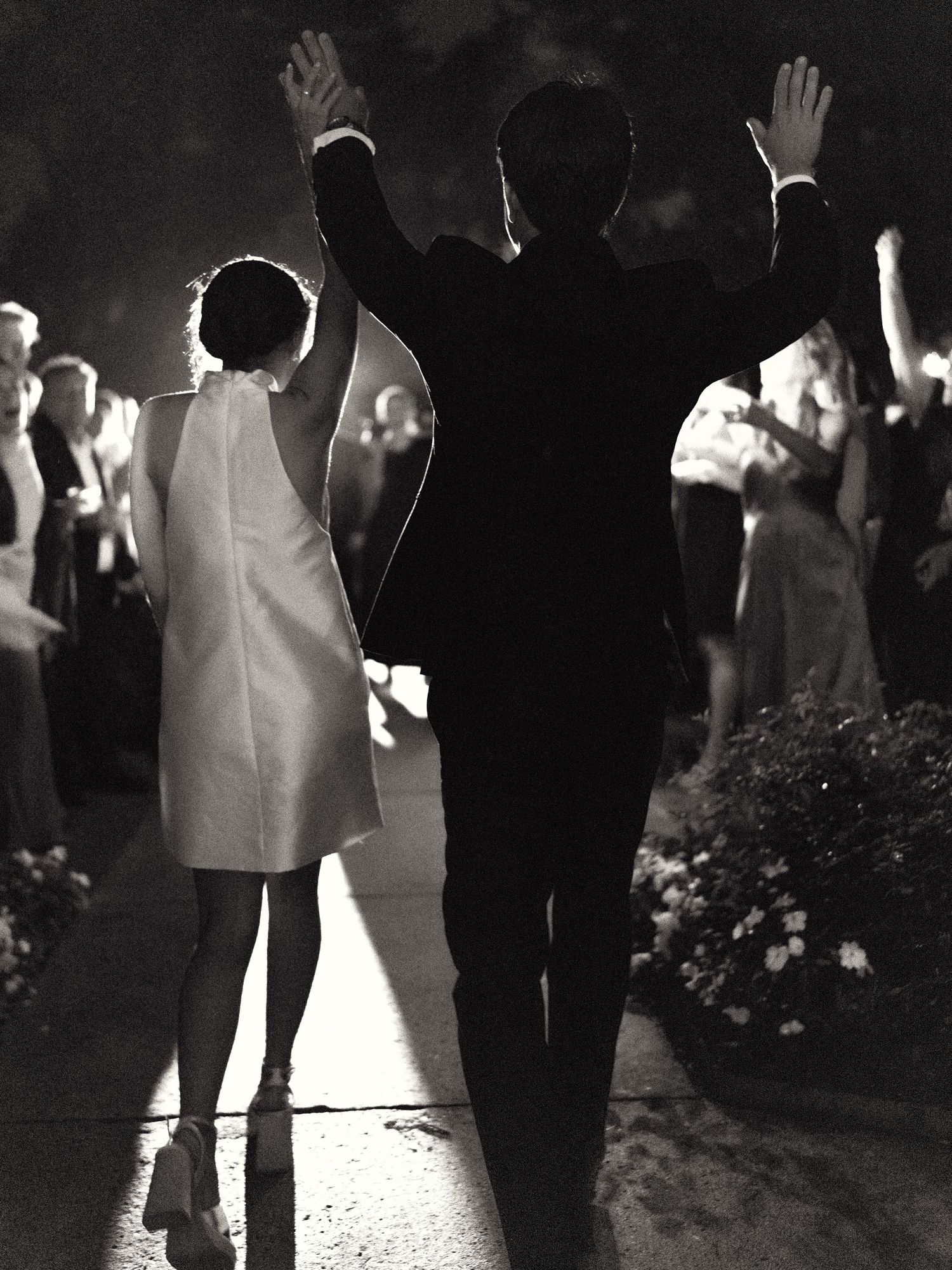 Black and white photo of bride and groom raising hands while walking through a cheering crowd.