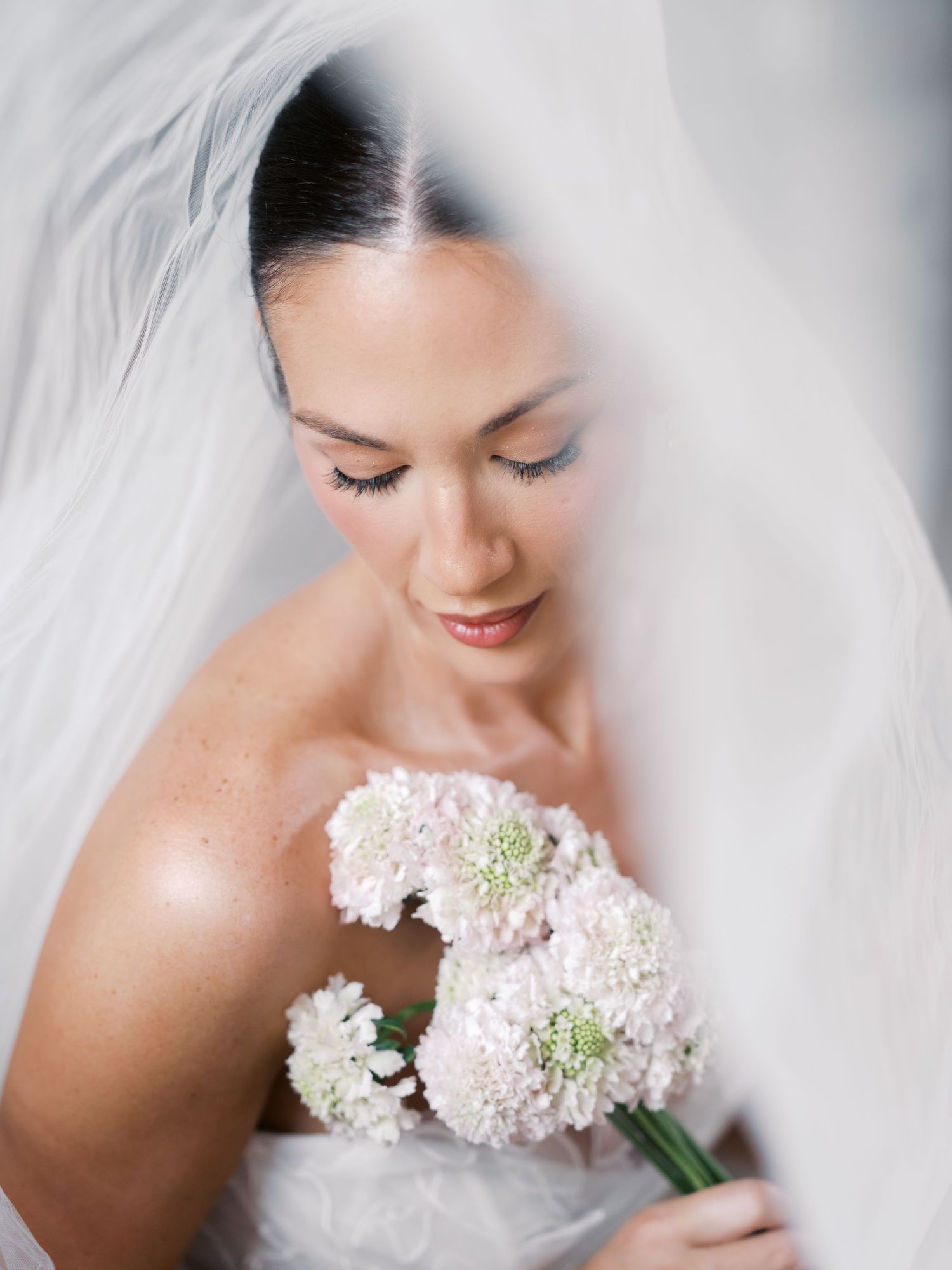 Serene bride with eyes closed holding soft pink scabiosa bouquet under flowing veil.