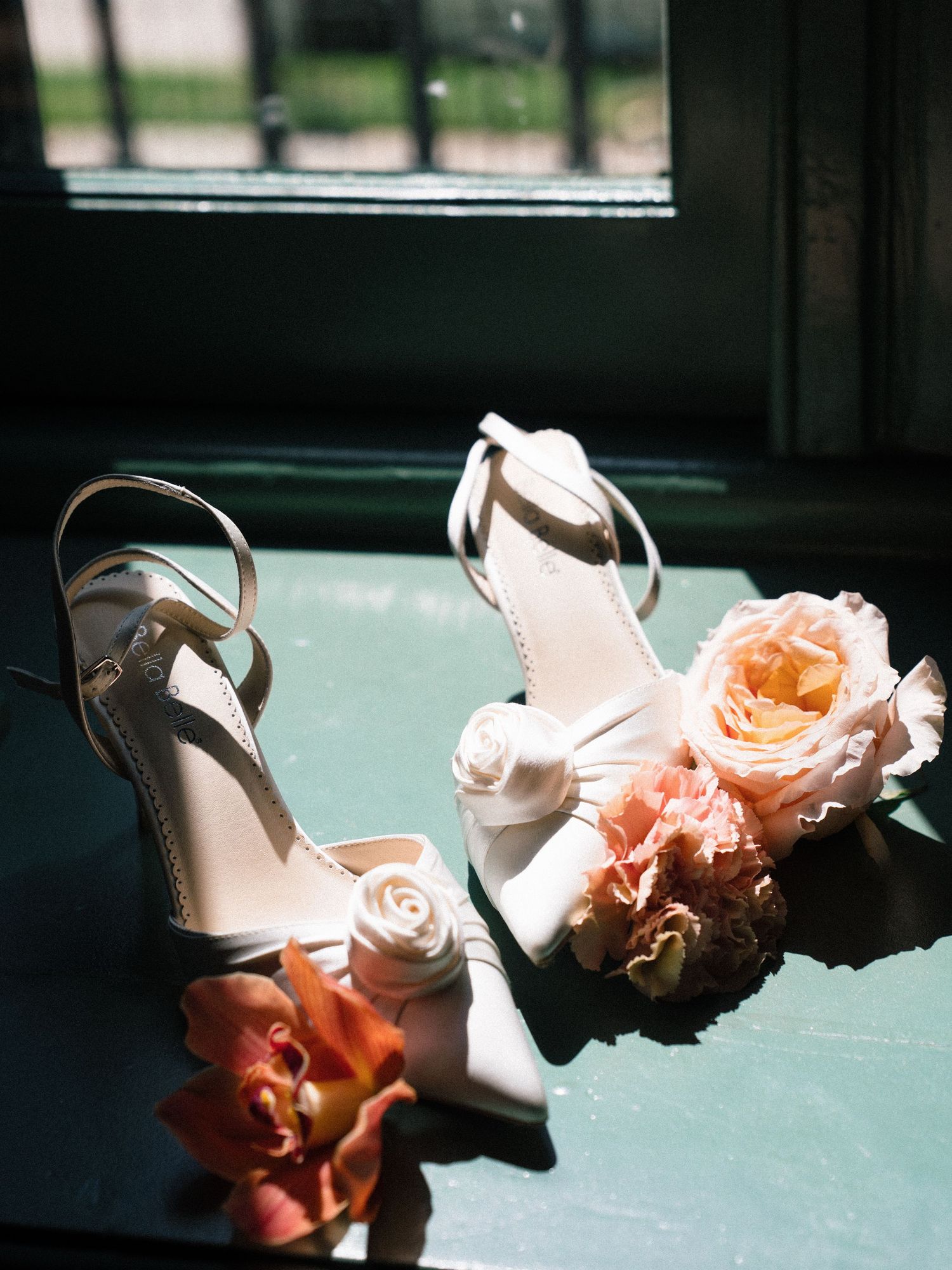 White bridal heels with rose detail surrounded by peach and coral flowers on a teal surface.