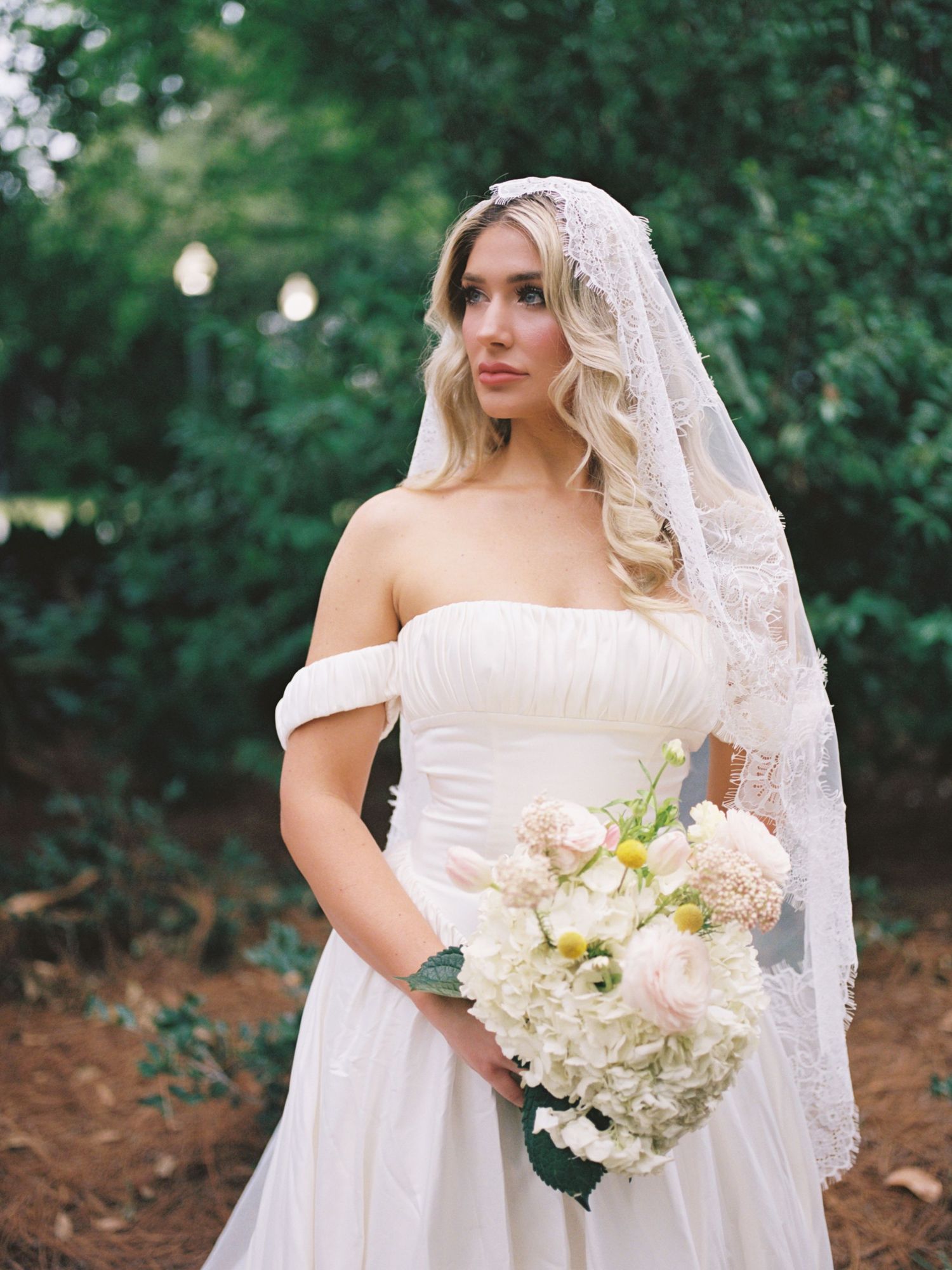 Bride in off-shoulder white gown with lace veil holds blush bouquet outdoors.