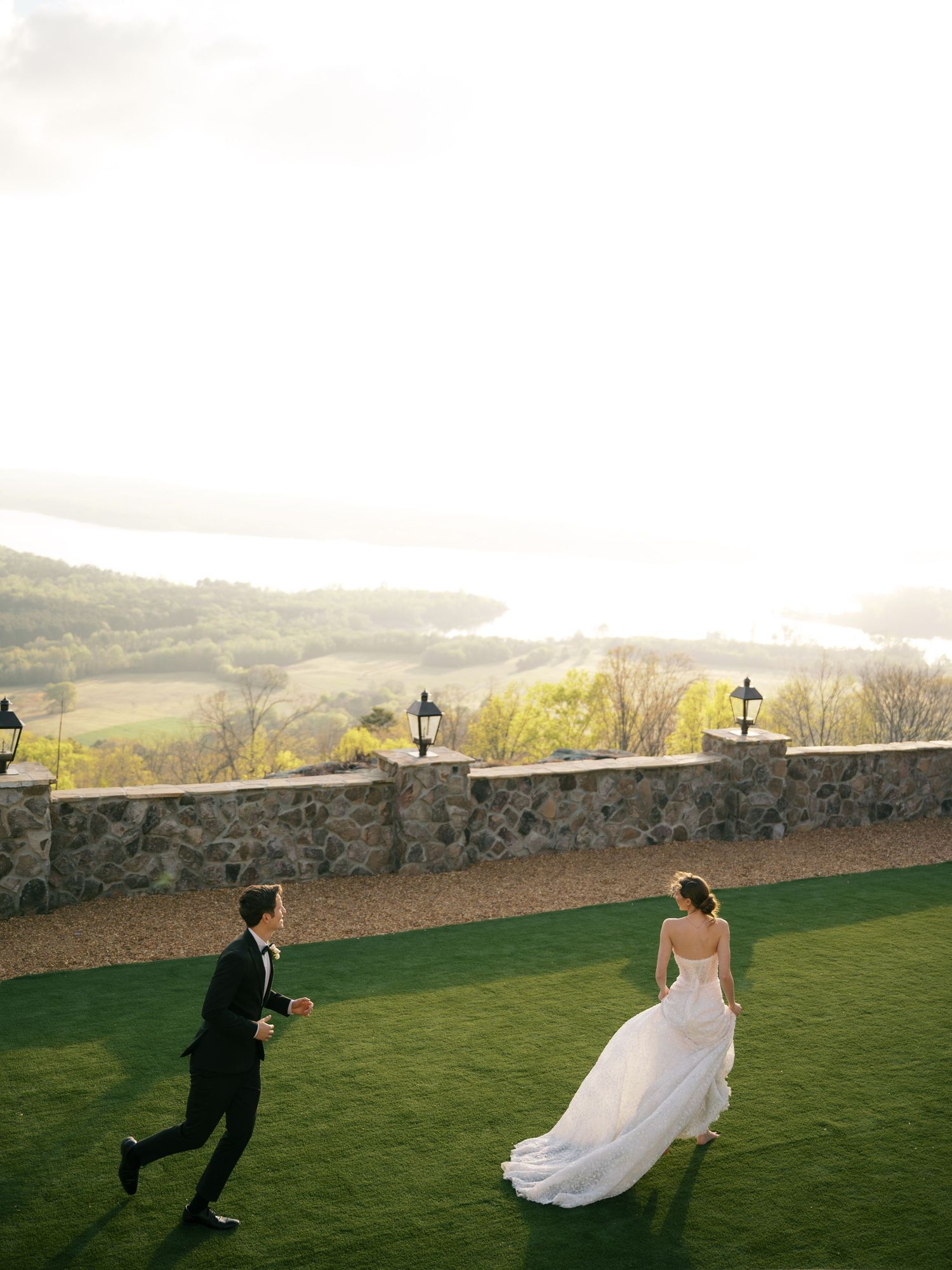 Bride in a flowing gown and groom running across a green lawn with a scenic valley view.