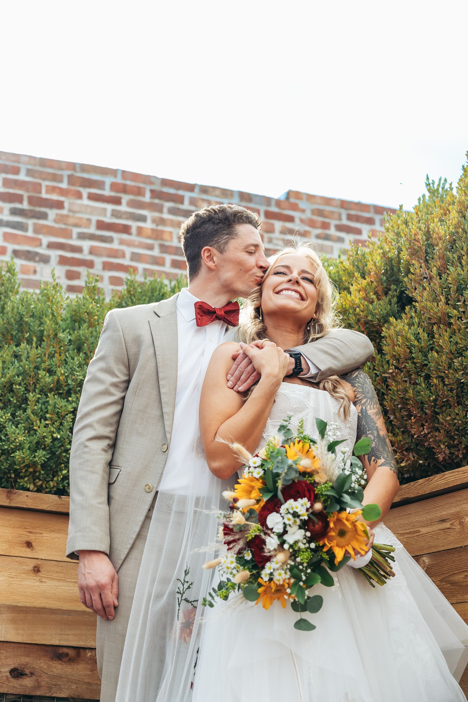 Groom in tan suit and red bow tie kisses smiling bride holding sunflower bouquet outdoors near brick wall.