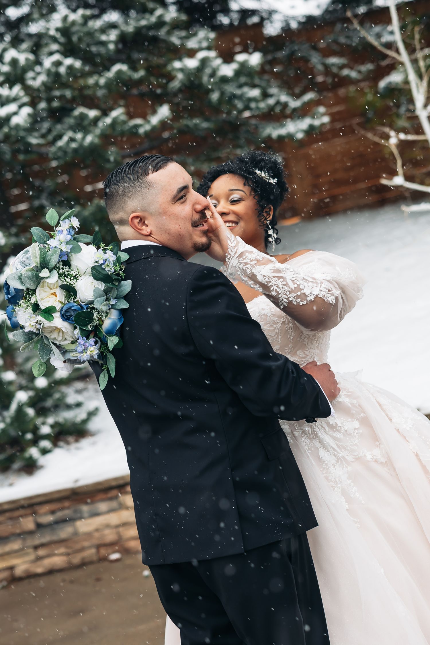 Bride and groom embrace in snowy winter wedding, bride in lace gown holds groom's face while snow falls around them.