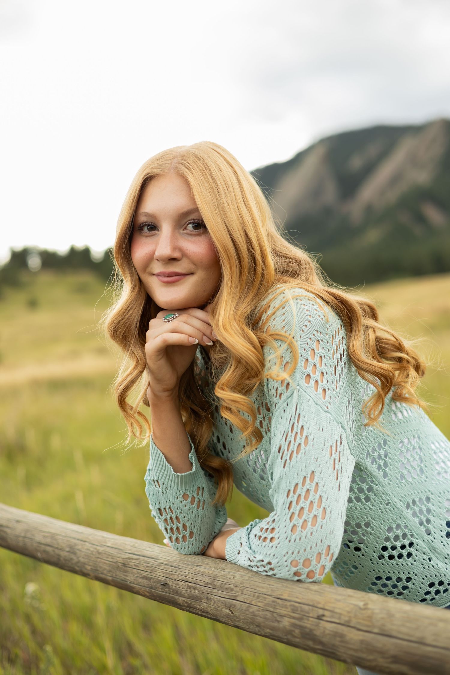 Portrait in mint lace shirt resting on wooden fence with mountain backdrop.