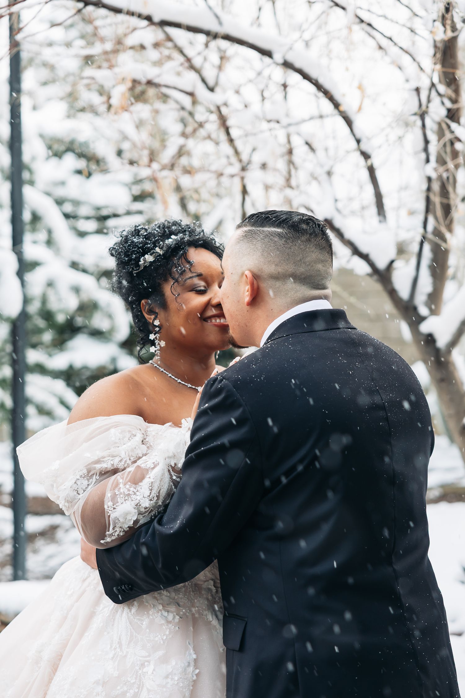 A bride and groom share a romantic kiss in a snowy winter wonderland, with the bride wearing an elegant off-shoulder lace gown.