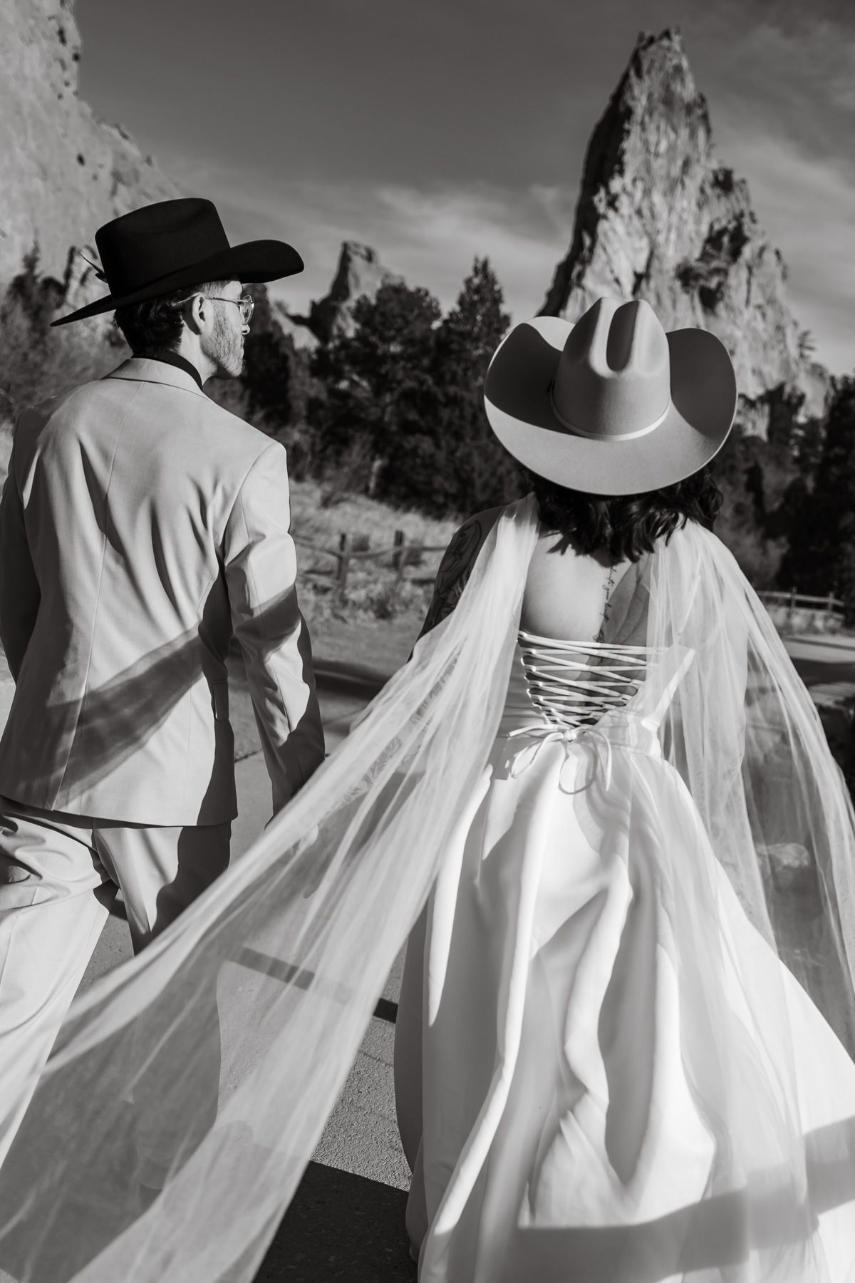 Black and white photo of a bride and groom in cowboy hats walking hand in hand with rocky formations in the background.