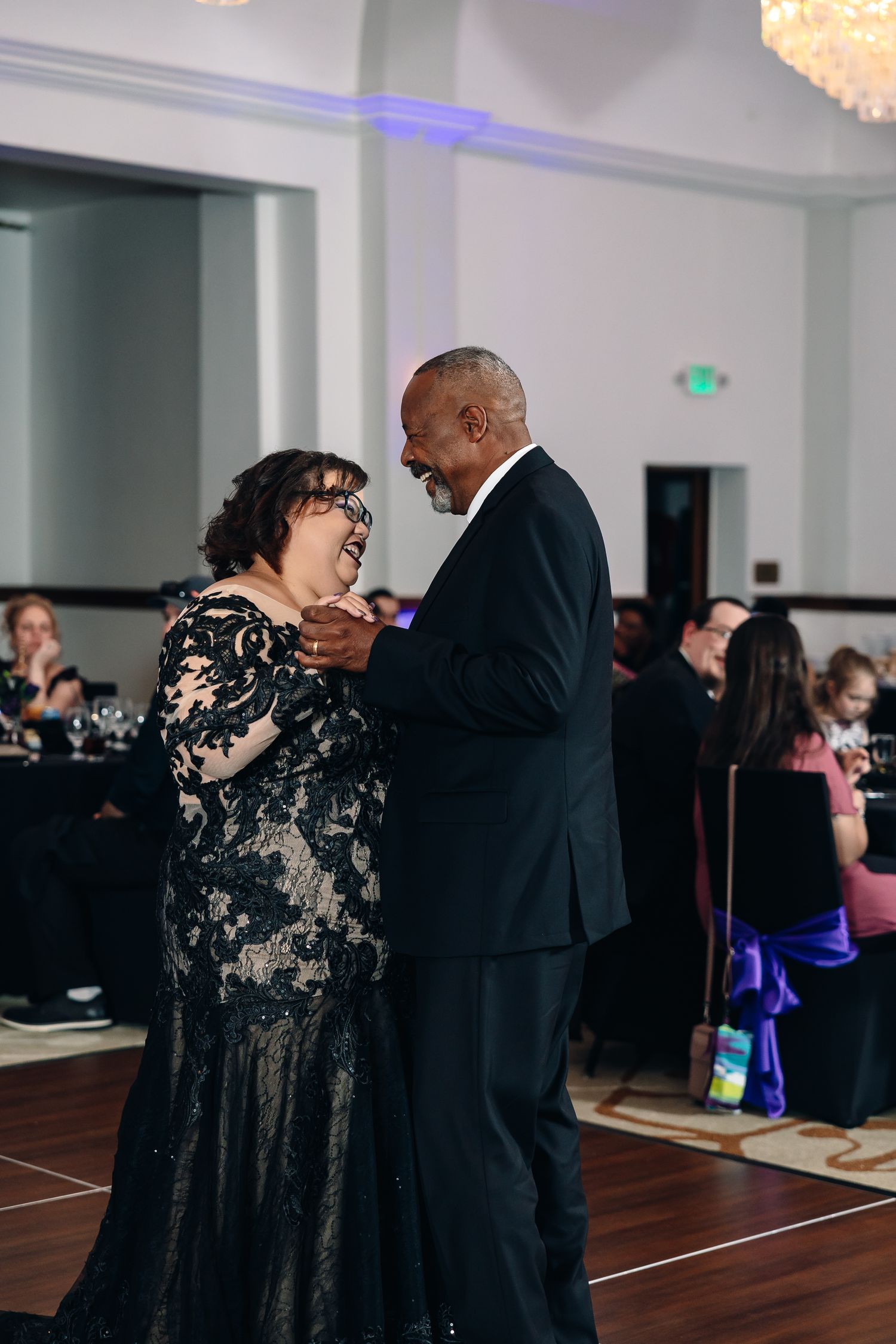 Elegant couple dancing at formal event, woman in black lace gown, man in suit, ballroom reception in background.