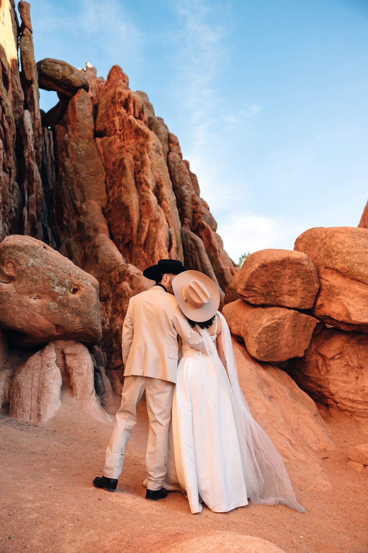 Couple in wedding attire embracing among red rock formations in a desert landscape, bride wearing white gown and hat.