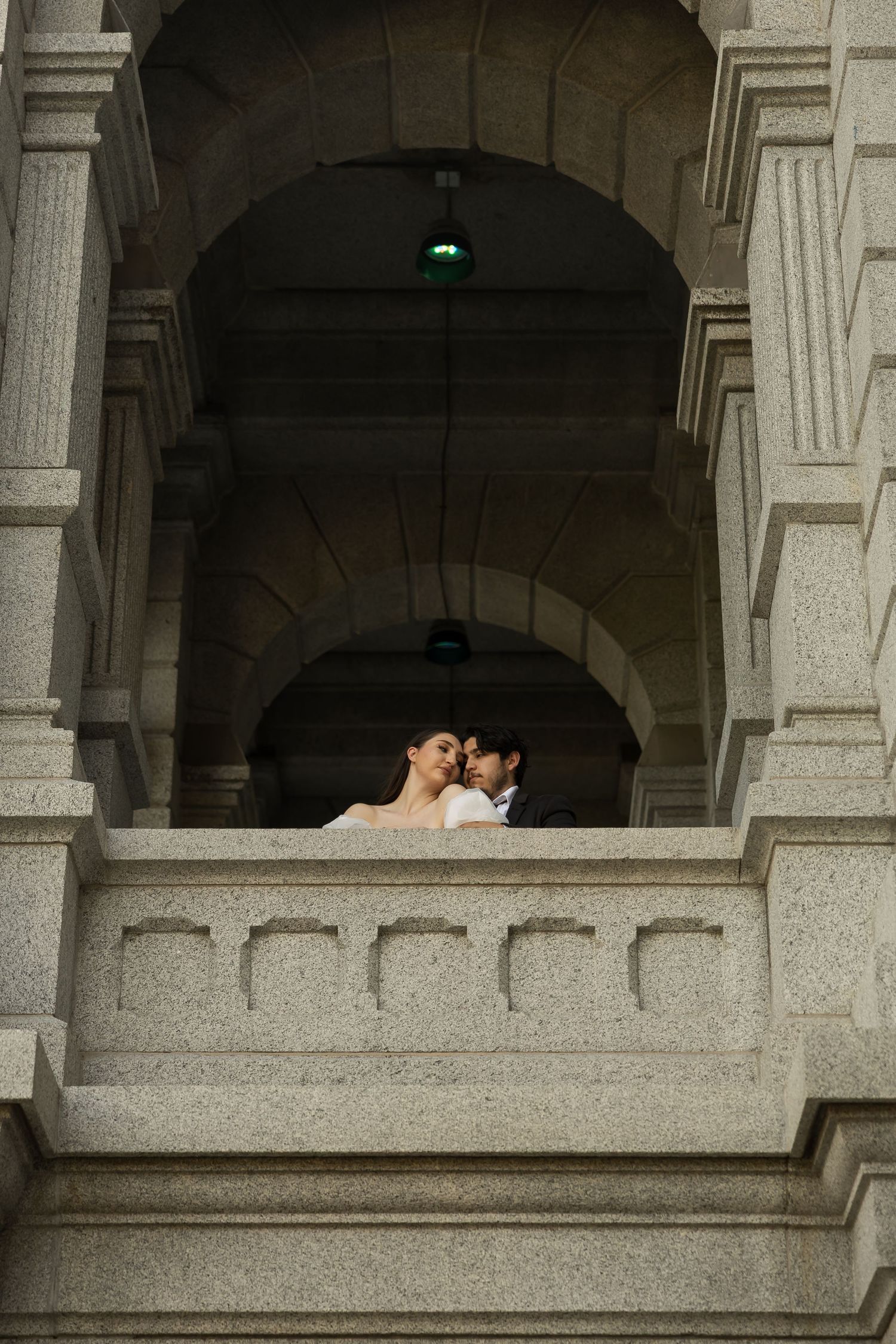 A wedding couple embraces in an arched stone balcony of a classical building.