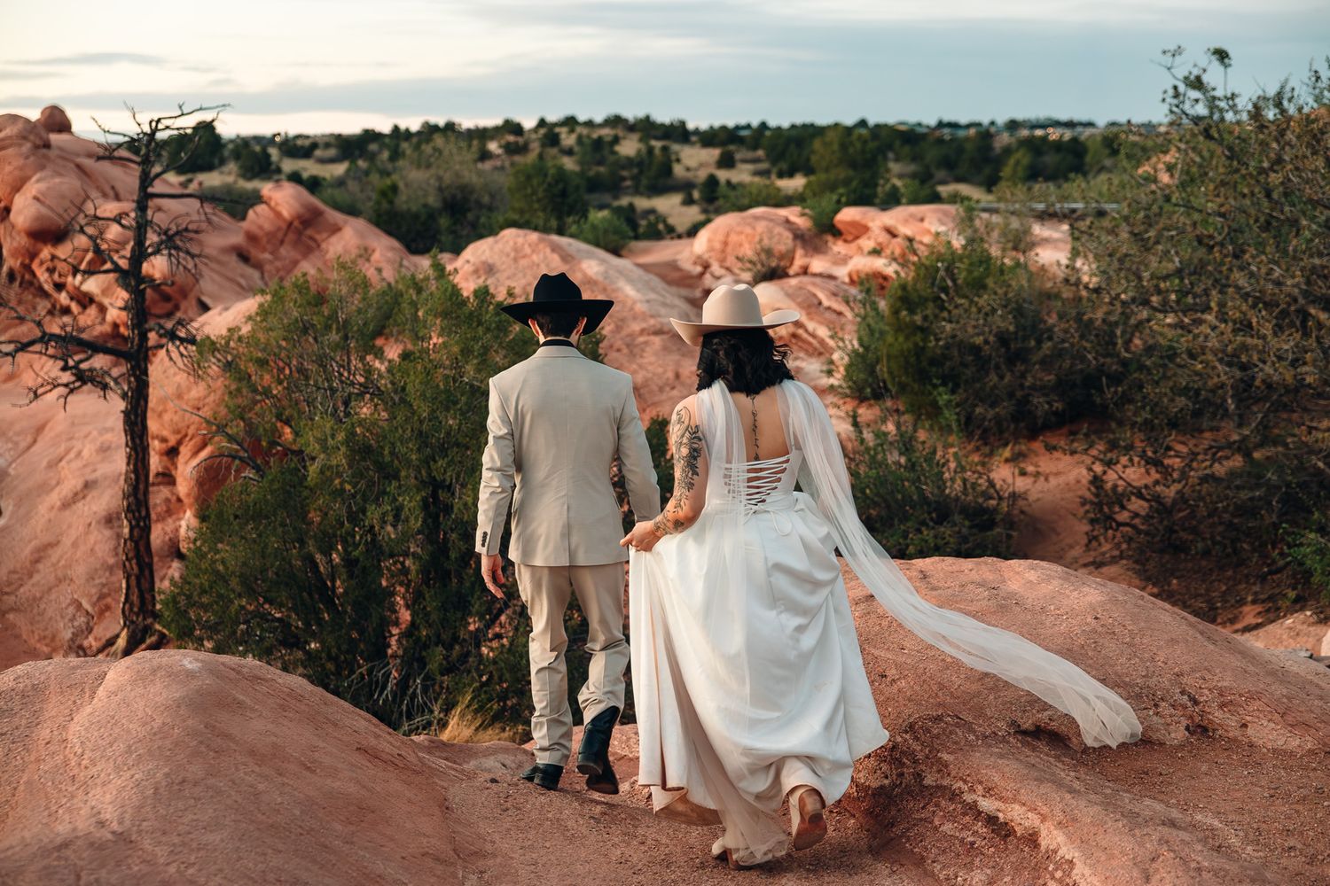 A bride and groom in western attire walk hand-in-hand through a stunning red rock desert landscape at dusk.
