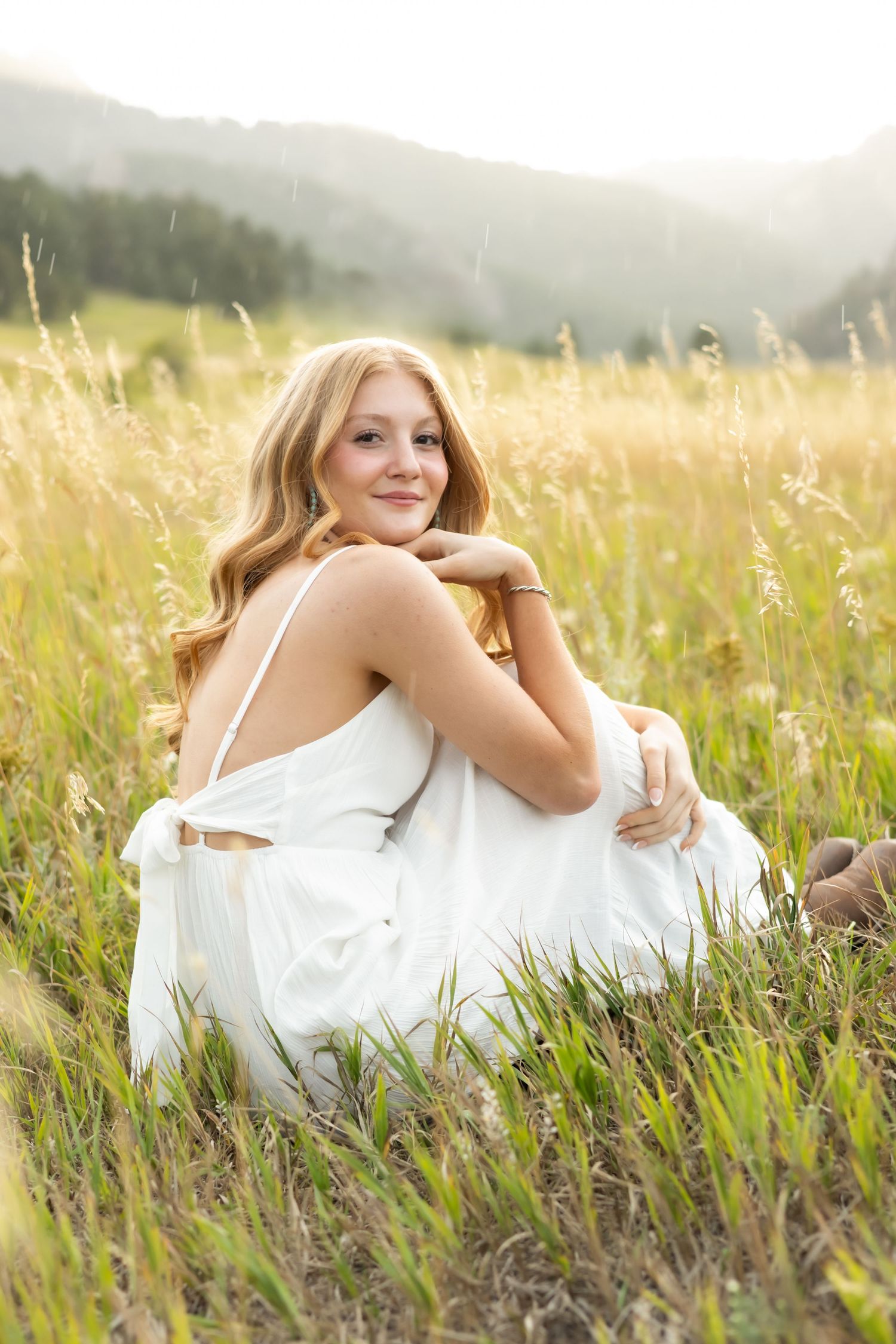 A white dress-clad figure sits gracefully in a golden meadow with misty mountains in the background.