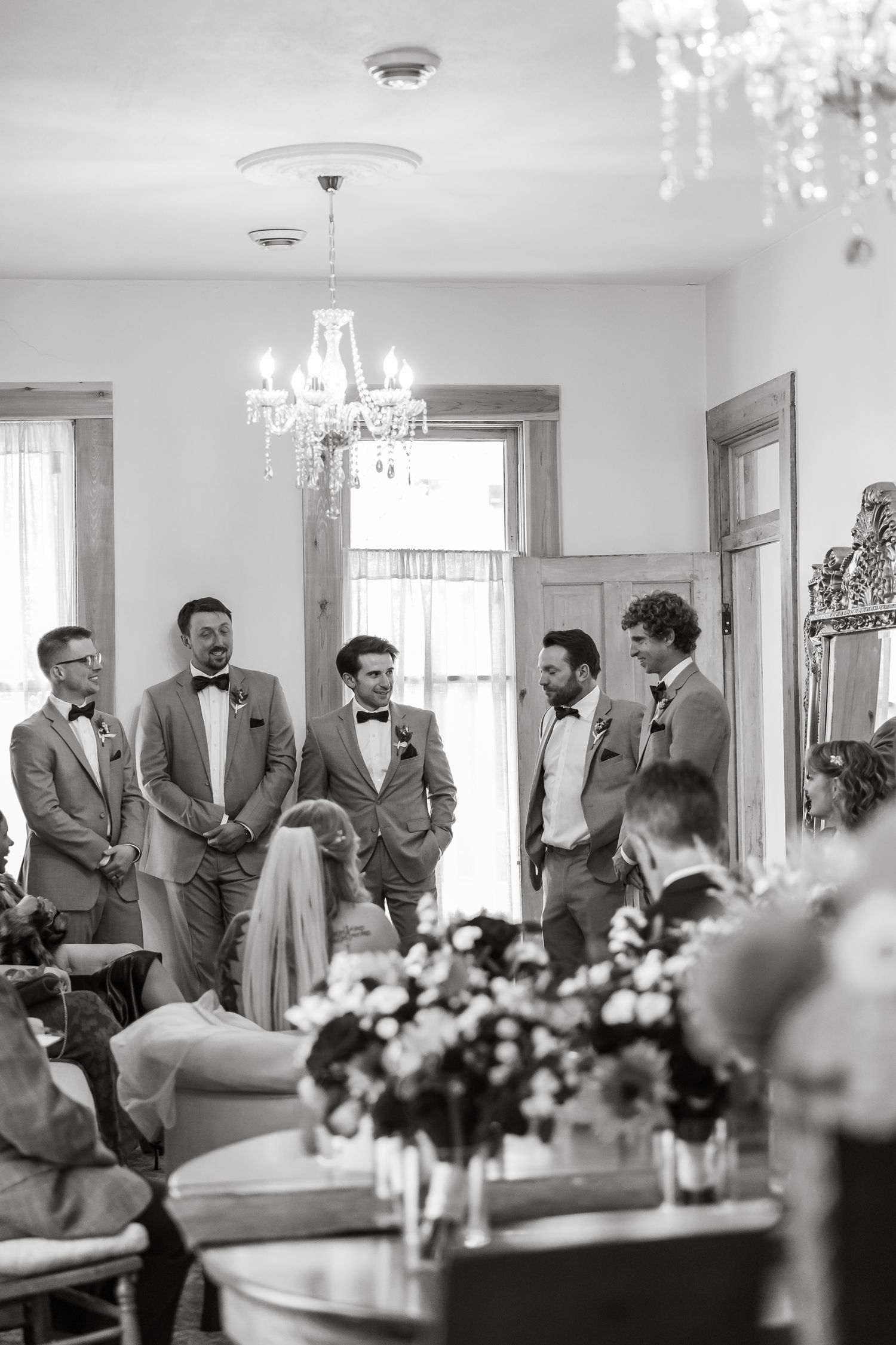 Black and white photo of groomsmen in suits gathered indoors at a wedding ceremony beneath an elegant chandelier.