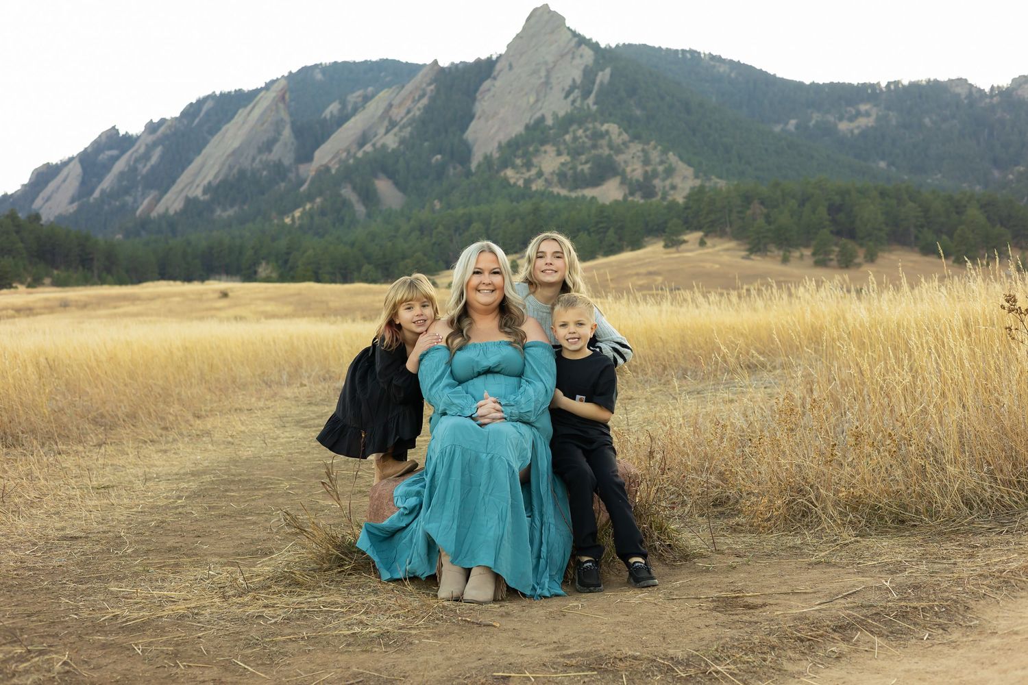 Pregnant woman in teal dress poses with three children in golden field with Flatirons mountains backdrop in Boulder, Colorado.