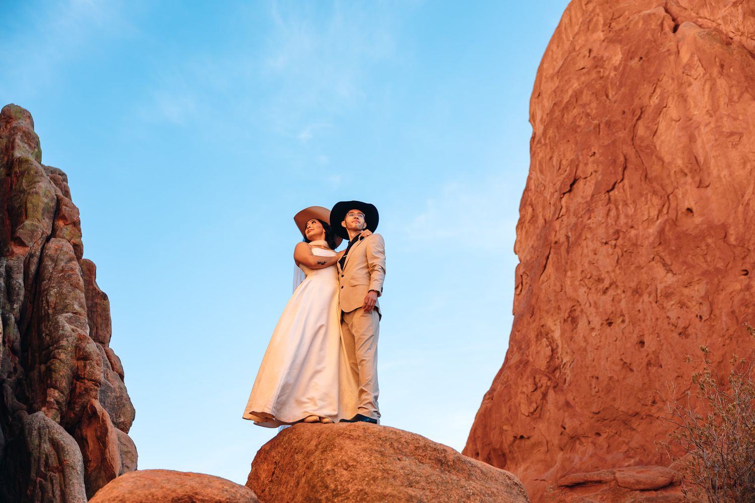 Couple in wedding attire kissing on red rock formations with blue sky backdrop at Garden of the Gods.