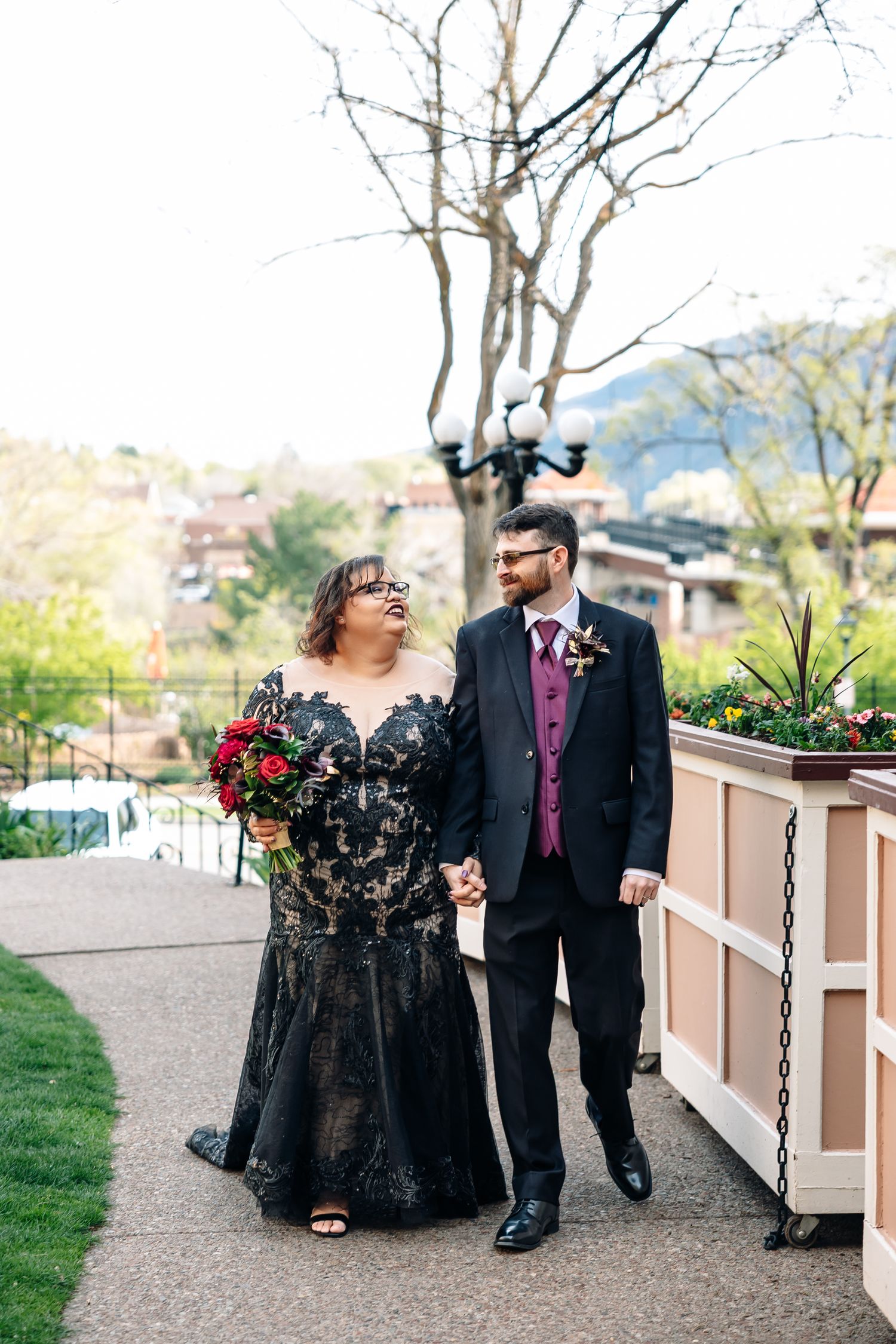 Couple walks hand-in-hand, bride in black lace gown with red bouquet, groom in black suit with pink vest.