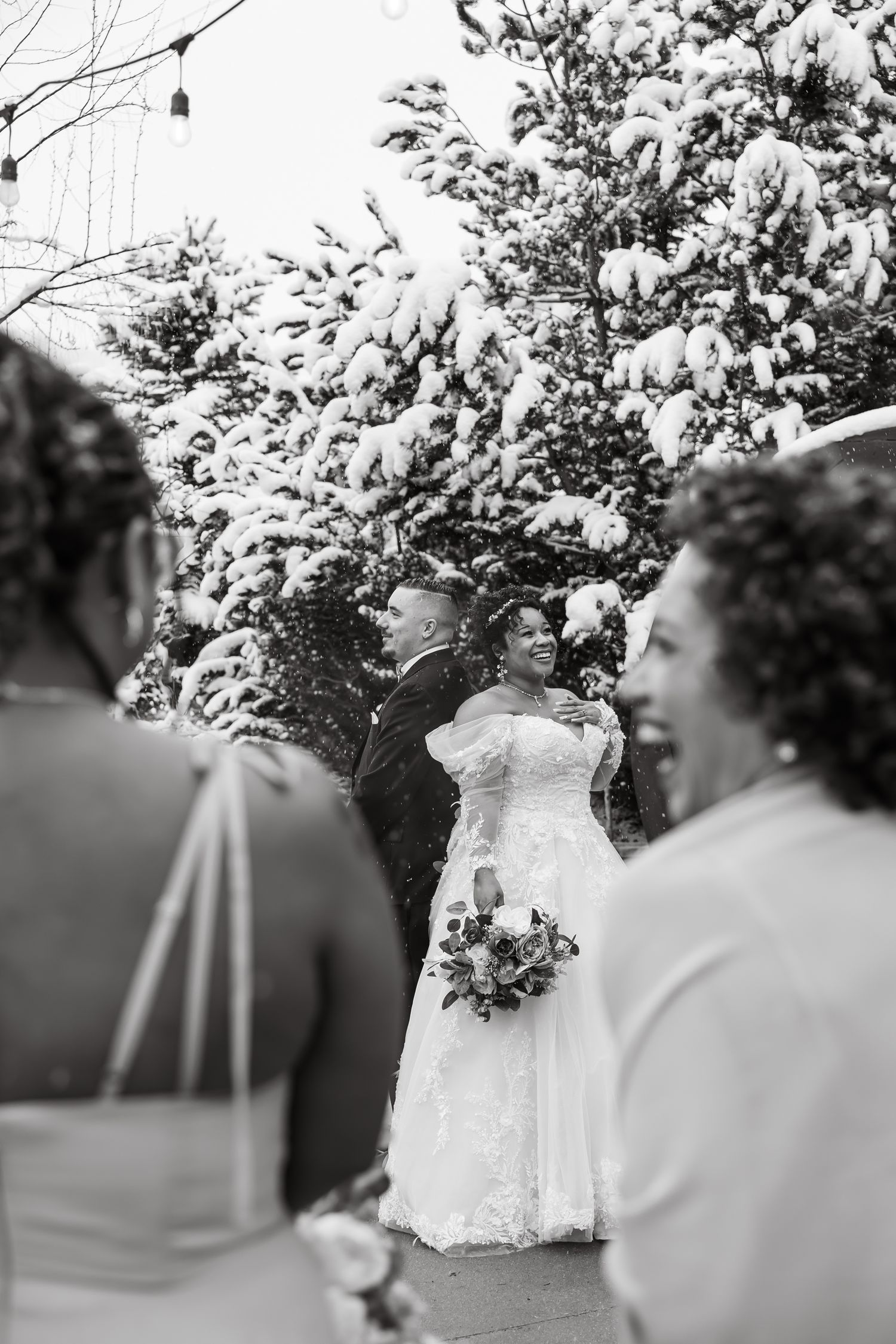 Black and white winter wedding ceremony with bride holding bouquet and groom standing before snow-covered evergreen trees.