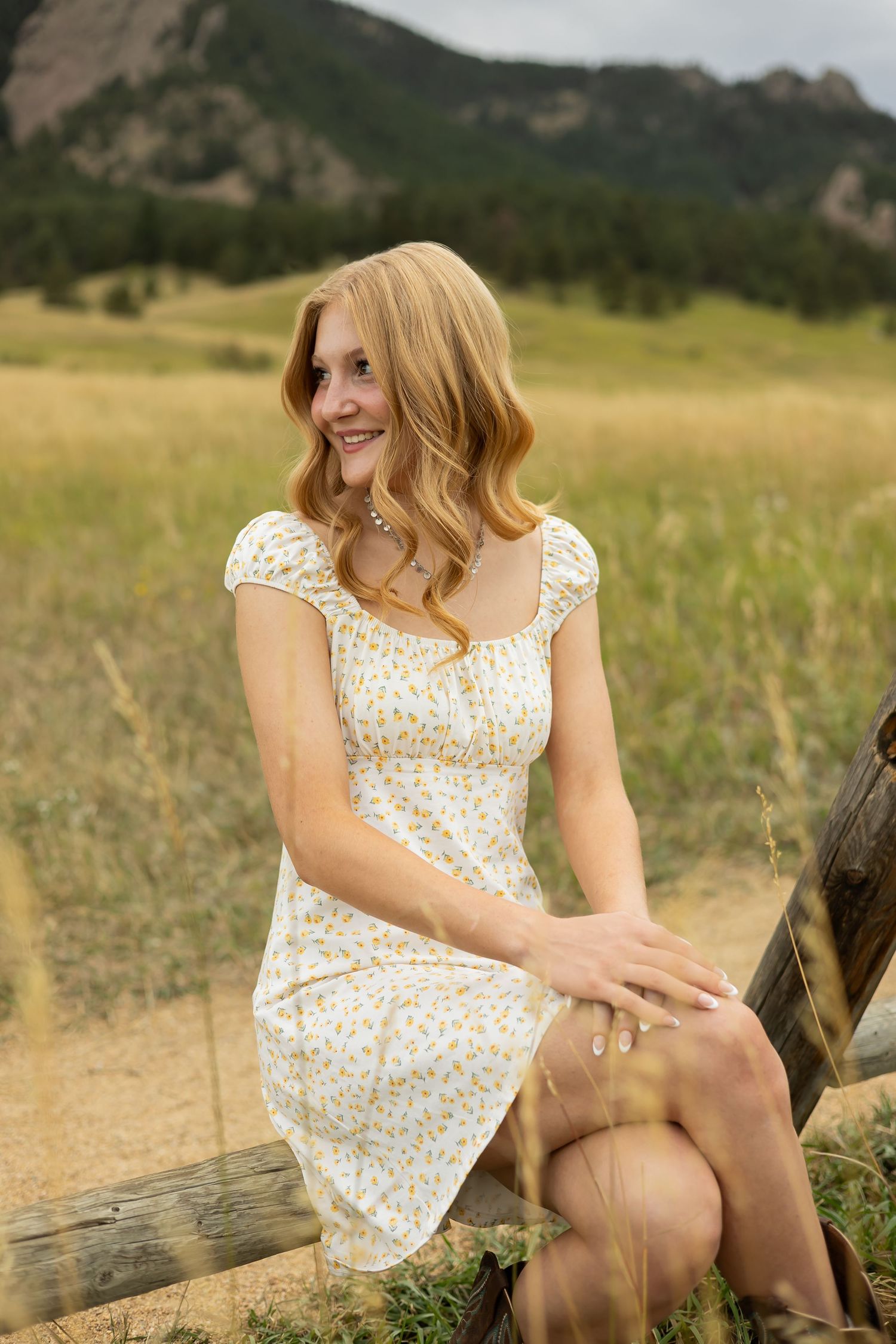Woman in floral dress sitting on rustic wooden fence with mountains in background.