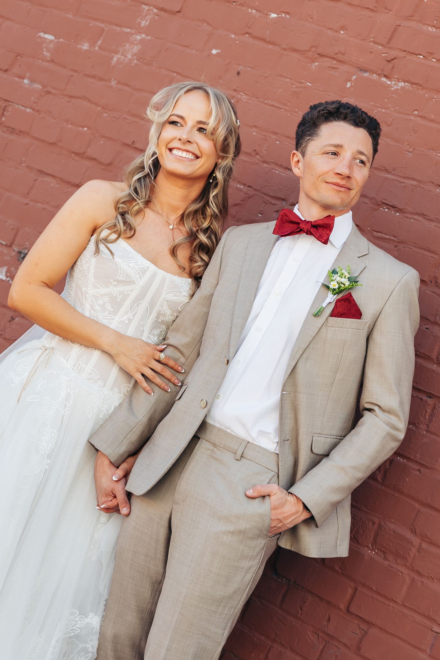 Smiling bride in white dress and groom in tan suit with red bow tie pose together against a brick wall.