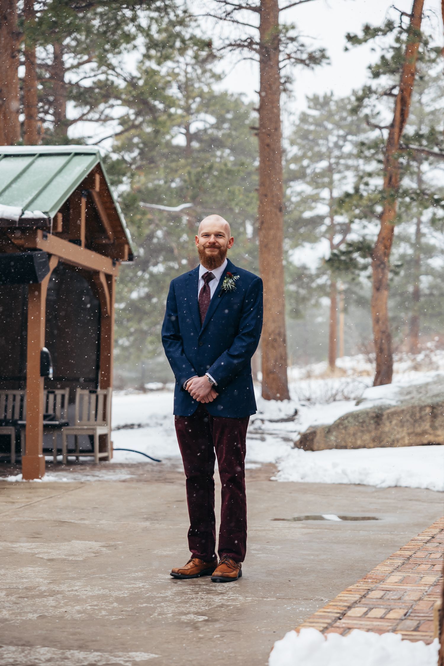 Groom in navy suit with burgundy tie stands in snowy winter wedding venue outdoor setting among pine trees.