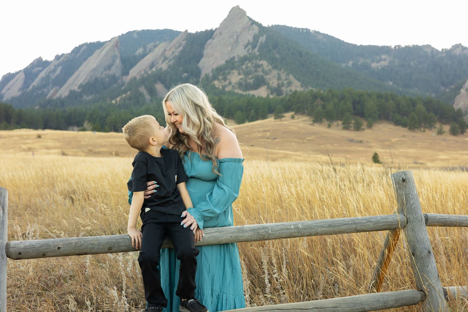 A mother and son share a tender moment on a wooden fence with the Flatirons mountains in the background.
