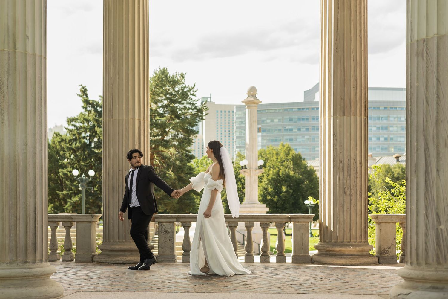 A wedding couple poses between classical columns with a scenic landscape view in the background.