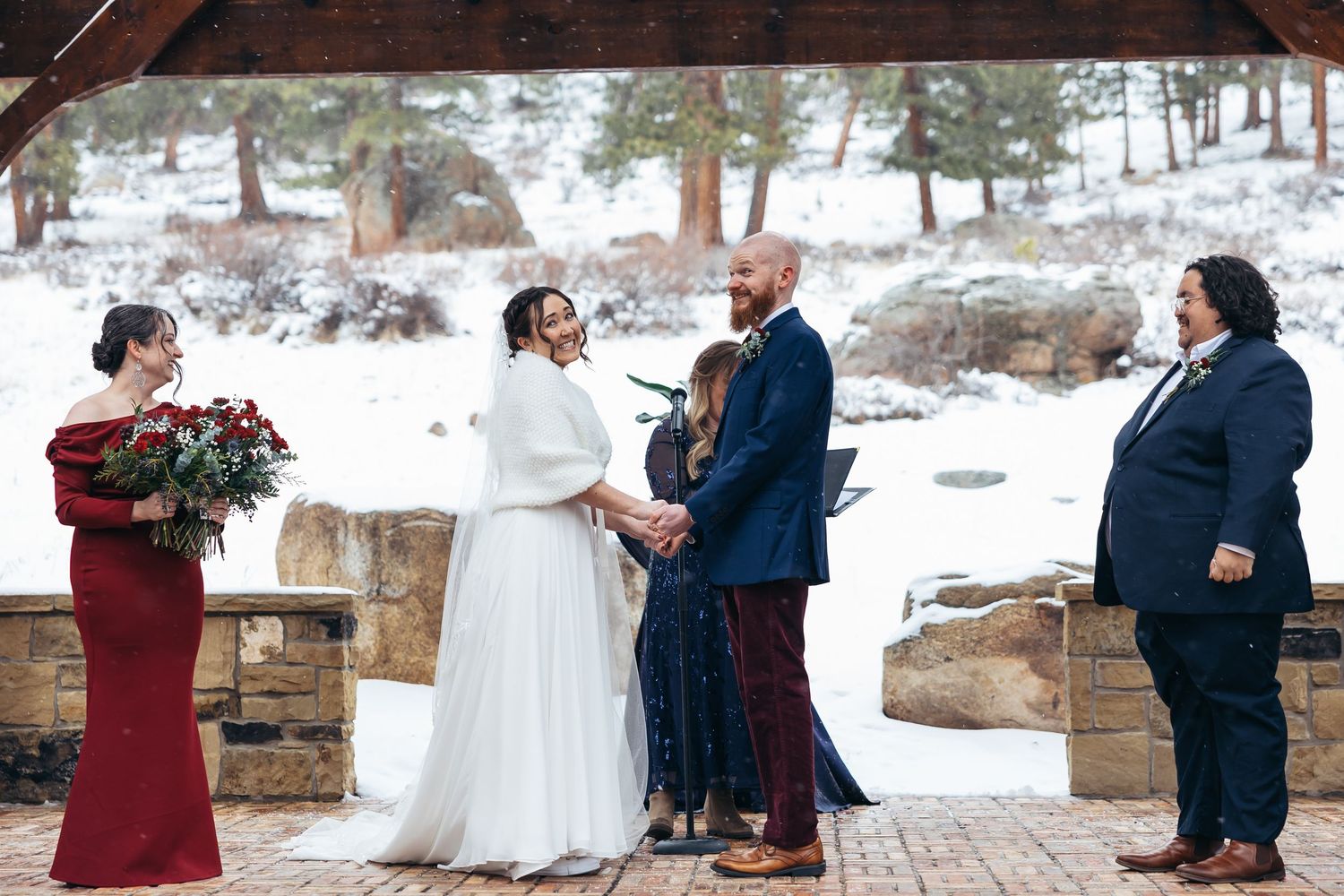 Snowy winter wedding ceremony outdoors with bride in white gown, groom in navy suit, and wedding party on stone patio.