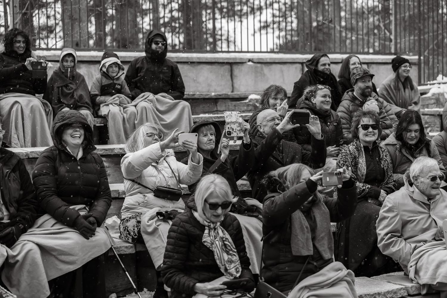 Black and white photo of elderly crowd seated on bleachers outdoors, bundled in winter coats, some taking photos.