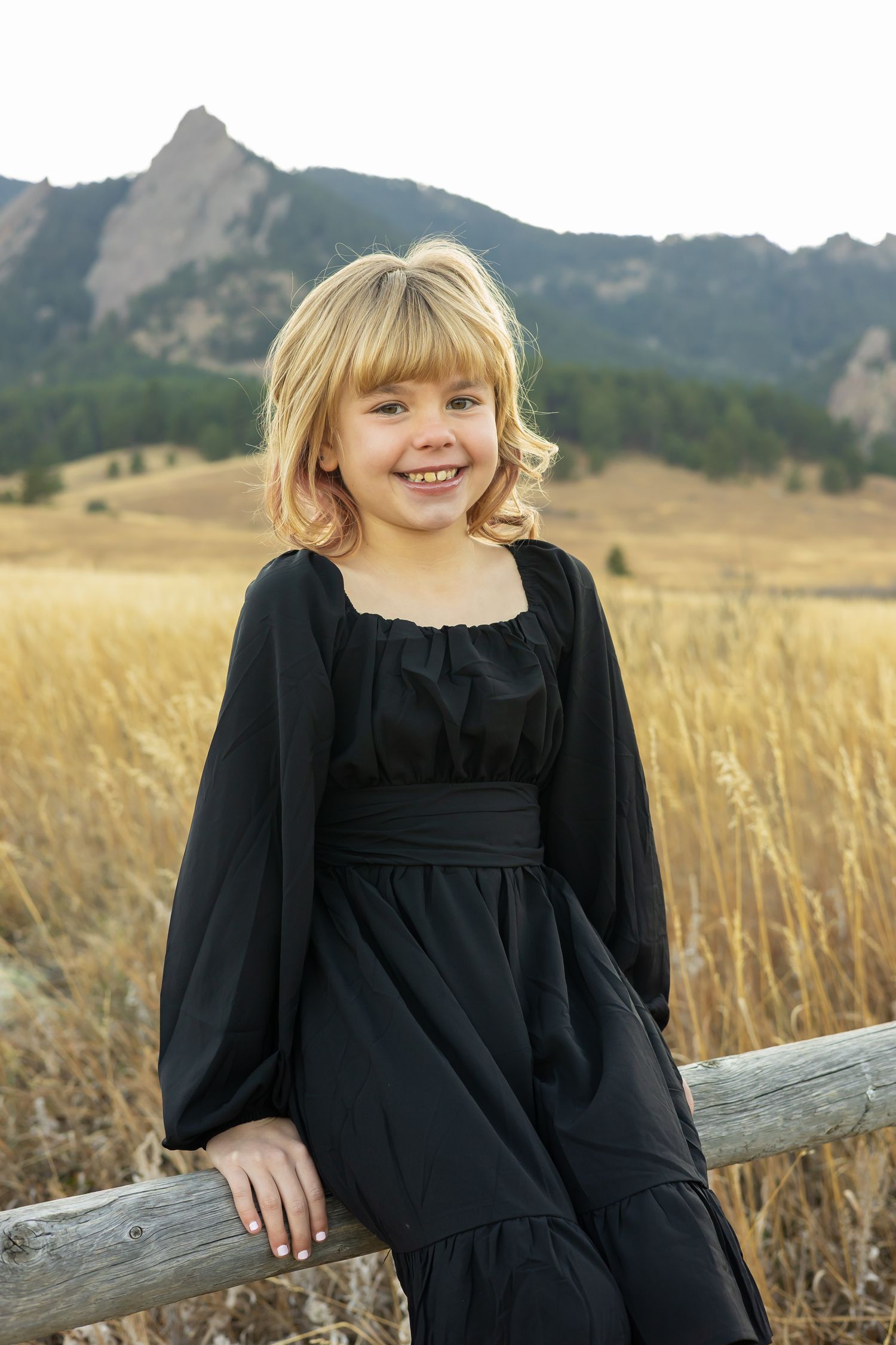 A child in a black dress sits on a wooden fence with the Boulder Flatirons mountains behind.
