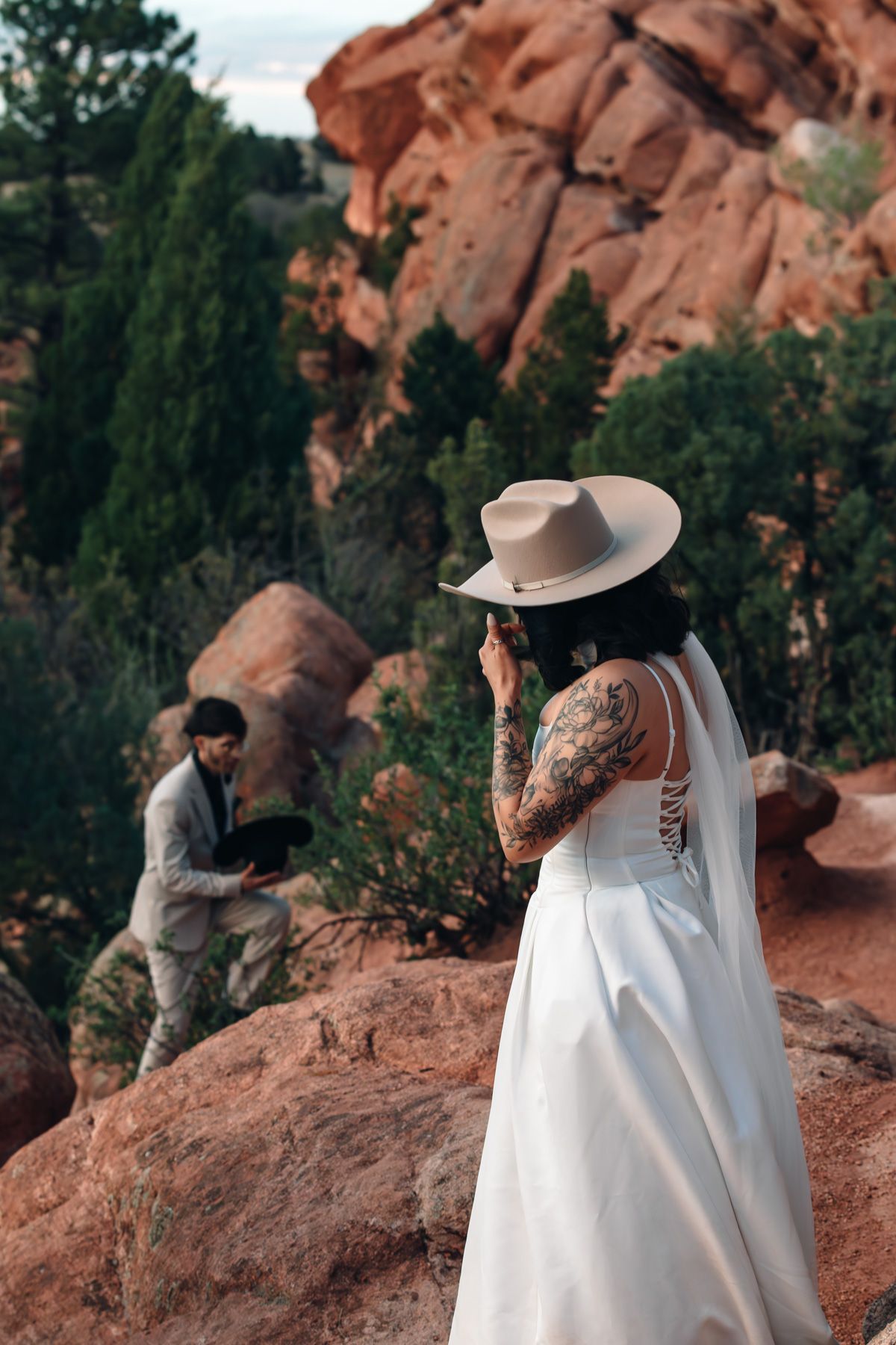 Tattooed bride in white gown and wide-brim hat stands on red rocks as groom kneels nearby at Garden of the Gods.