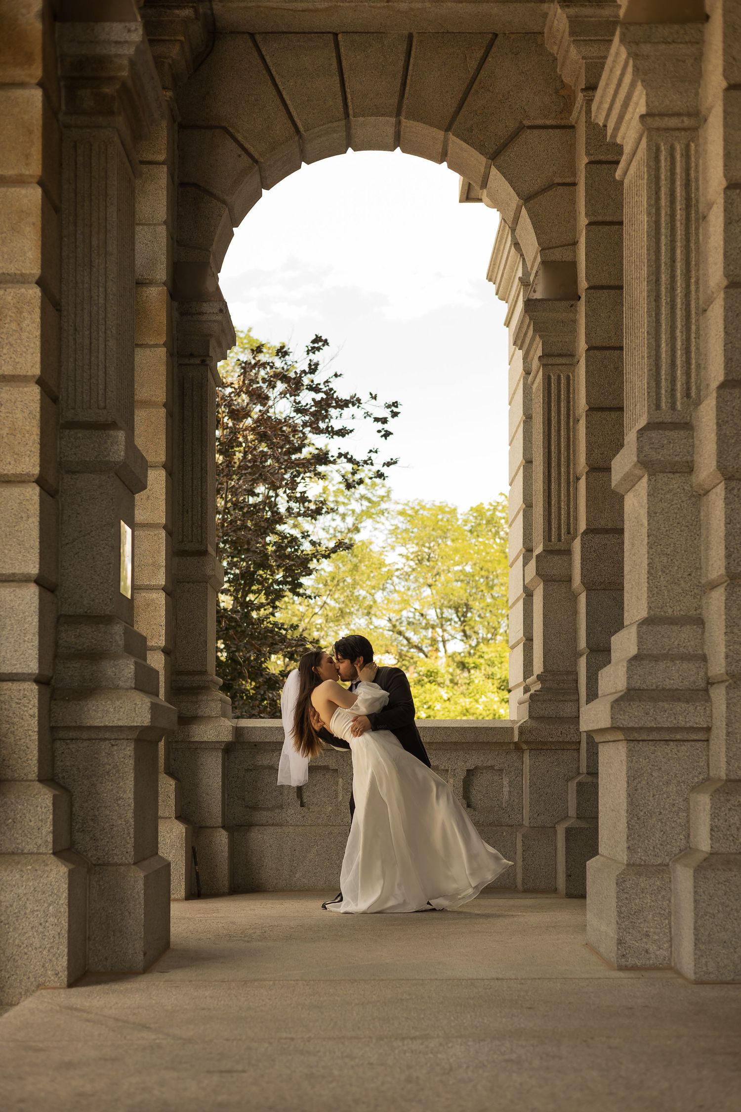 Wedding couple photographed in arched stone colonnade with natural lighting.