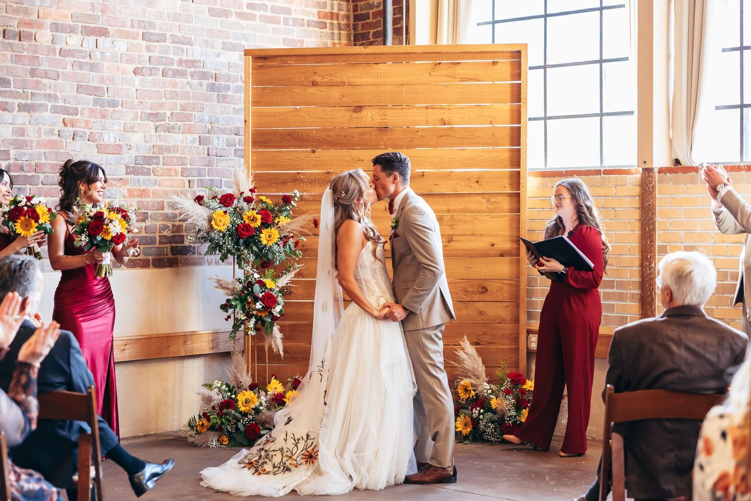 Bride and groom share first kiss at rustic indoor wedding ceremony with floral arrangements and brick walls.