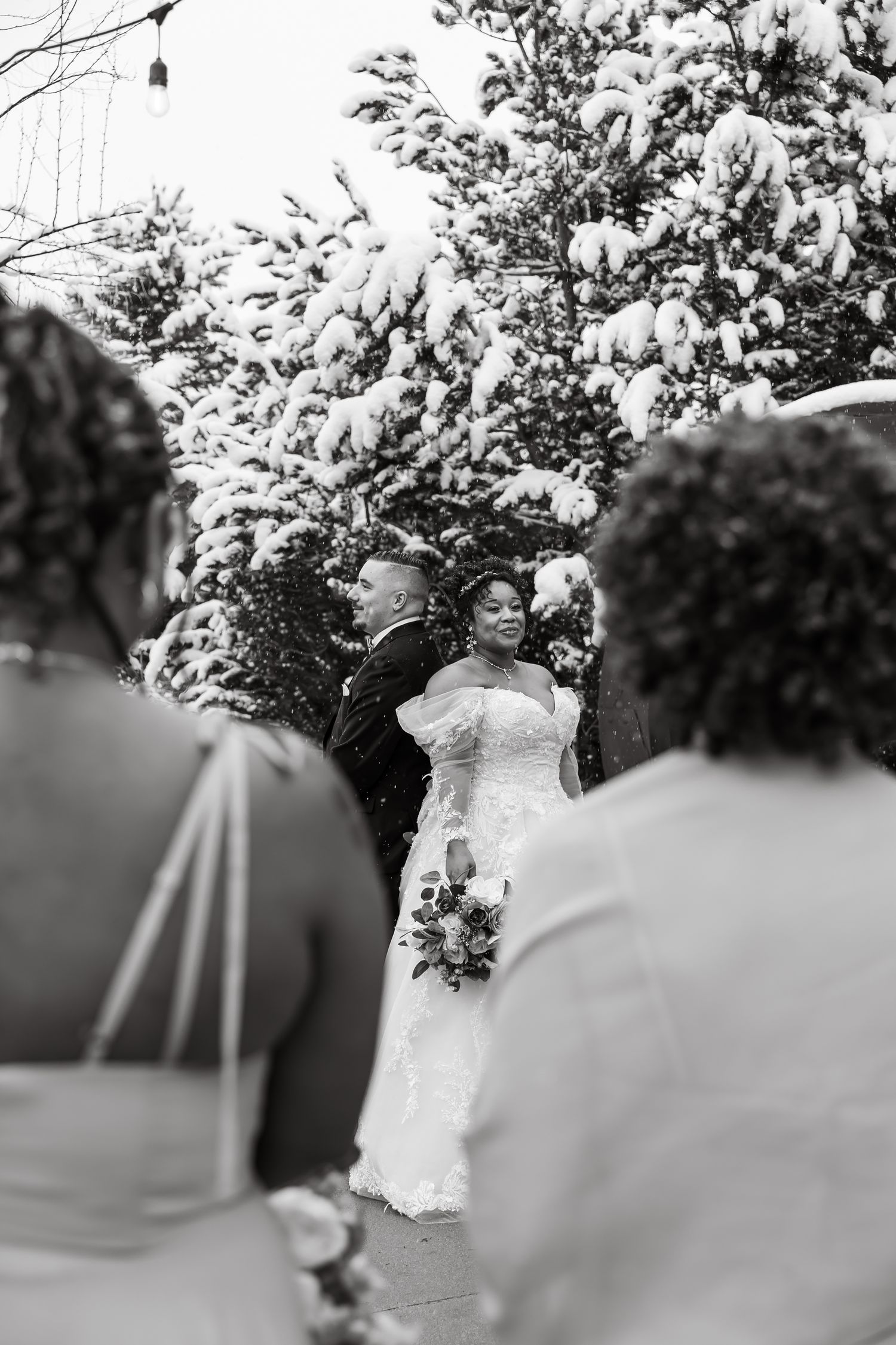 Black and white winter wedding ceremony outdoors with snow-covered trees, bride in lace gown holding bouquet beside groom.