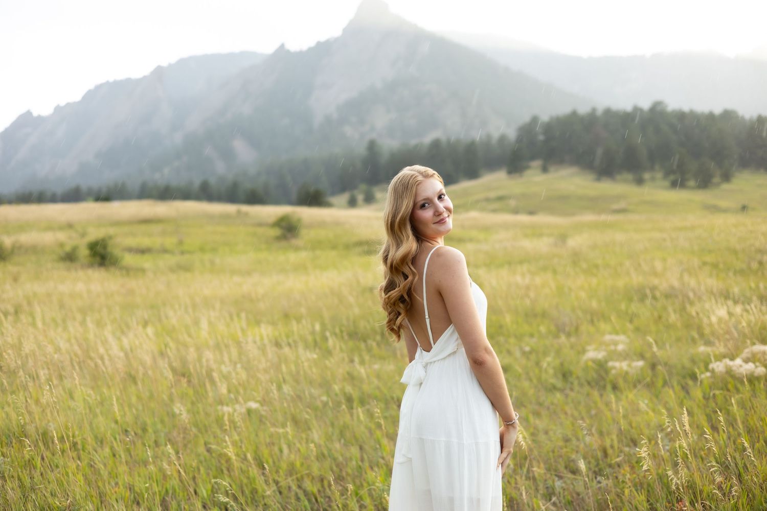 Woman in white dress smiling in a lush green meadow with the Flatirons mountains in the background.