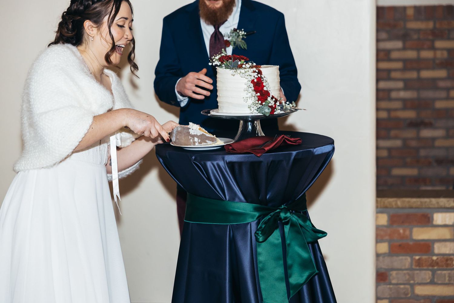 Bride and groom cutting a small white wedding cake decorated with red flowers on a navy blue draped table.