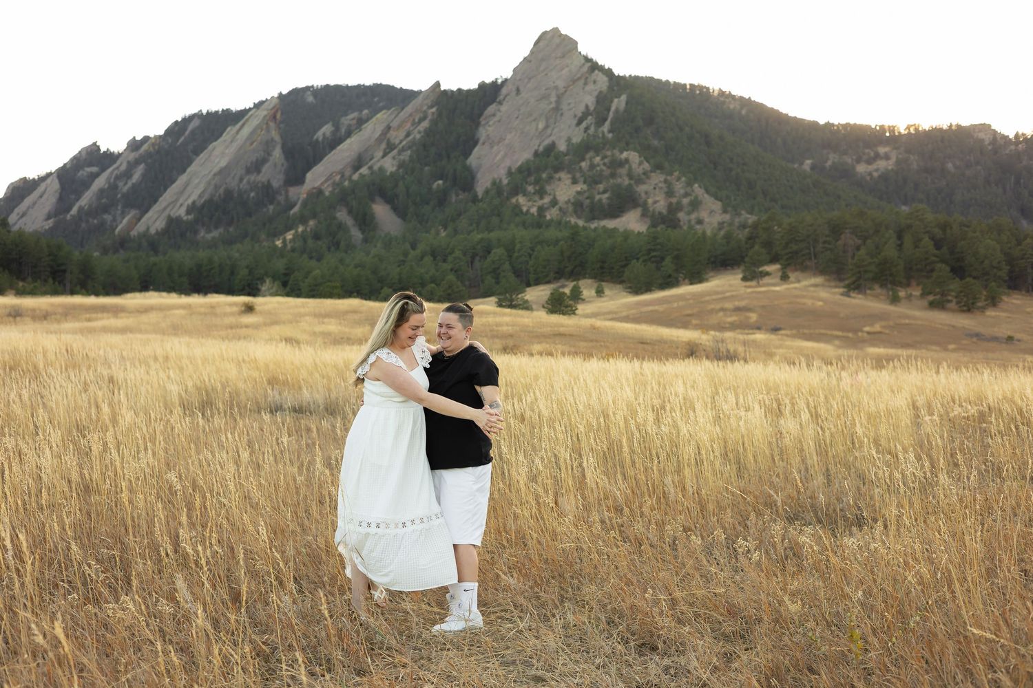 Couple embracing in golden field with Colorado Flatirons rocky mountains backdrop during sunset engagement photo session.