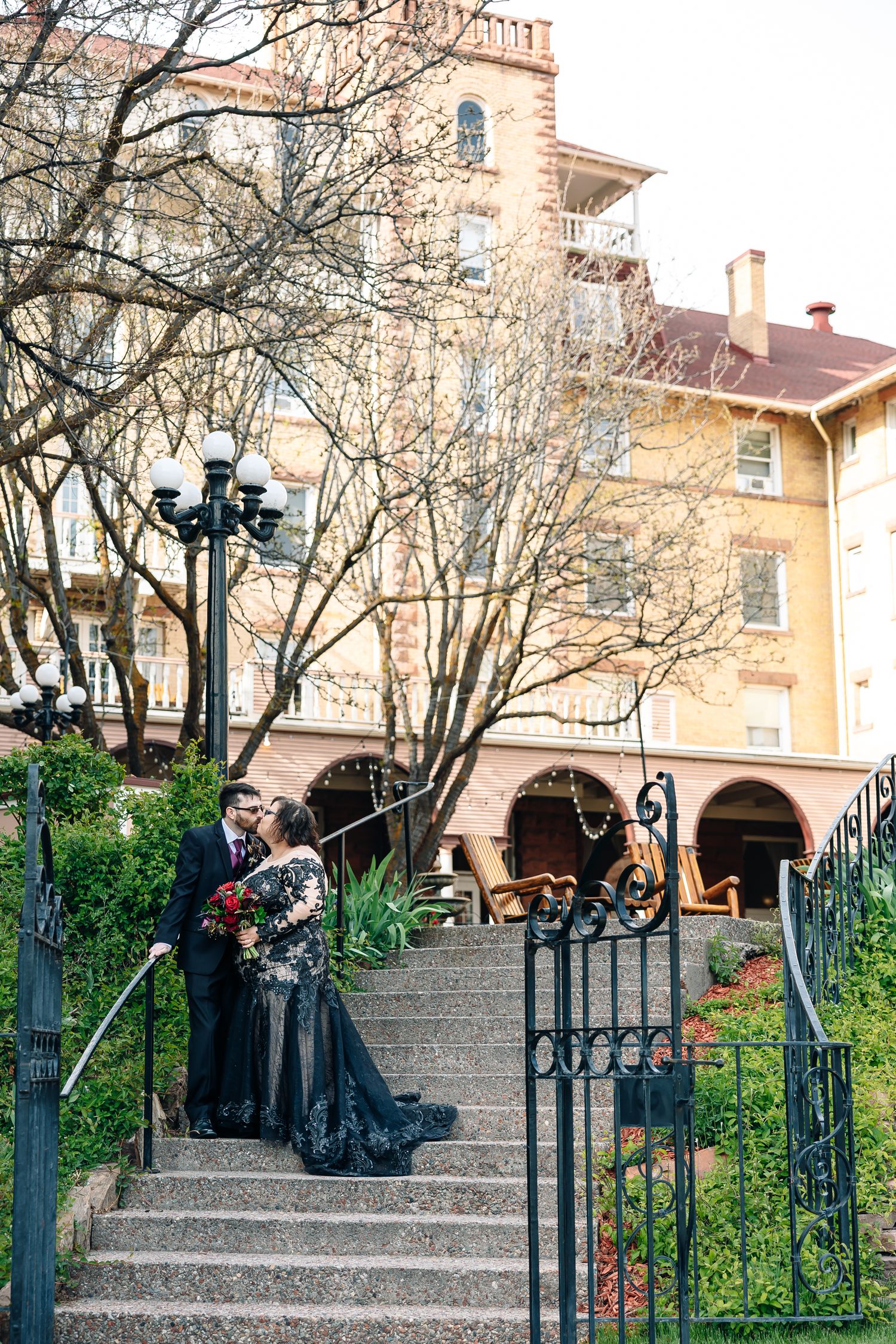 Couple in black wedding attire poses on stone steps outside a historic yellow building with iron railings and bare trees.