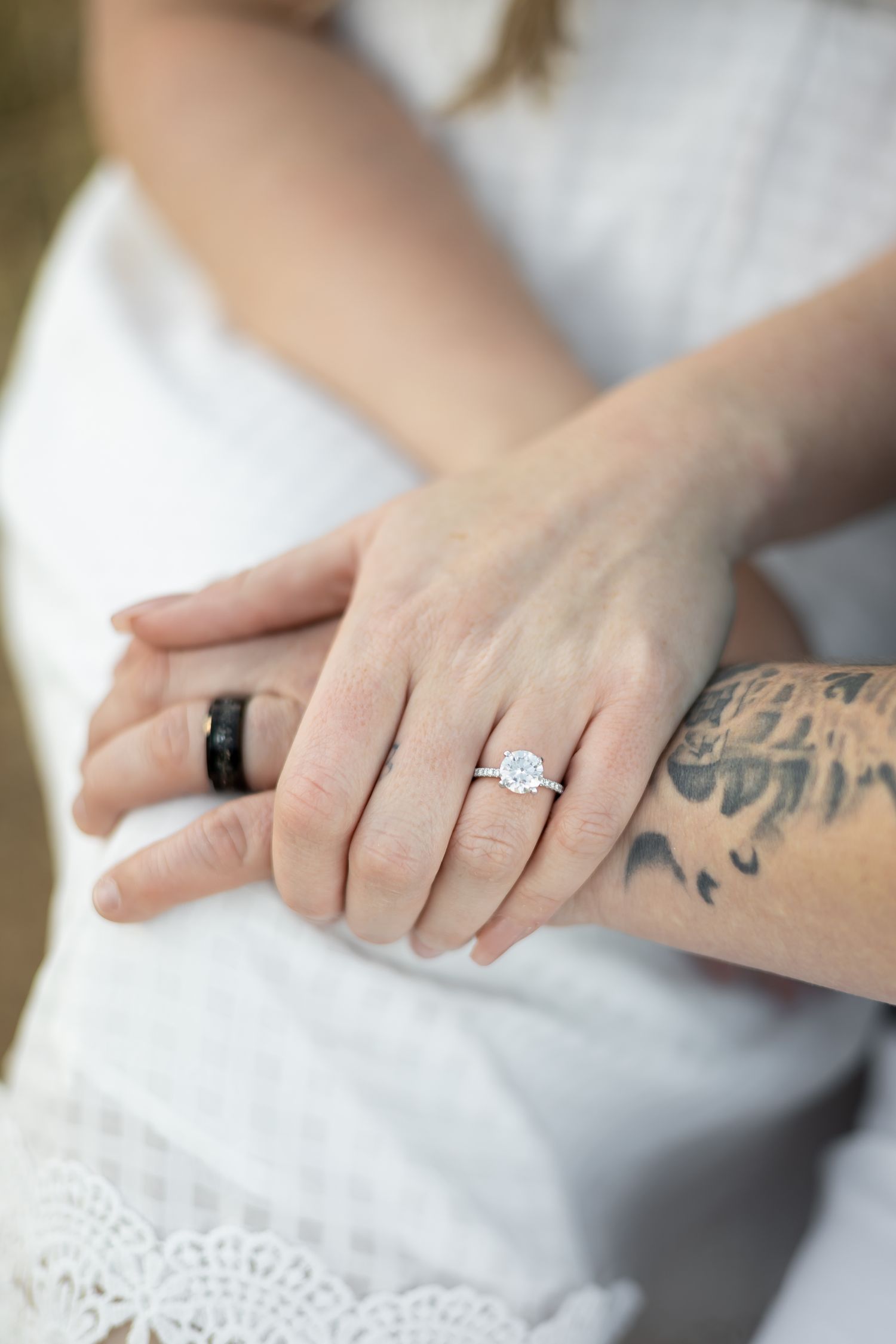 Close-up of hands wearing engagement ring and black wedding band with tattoos visible.