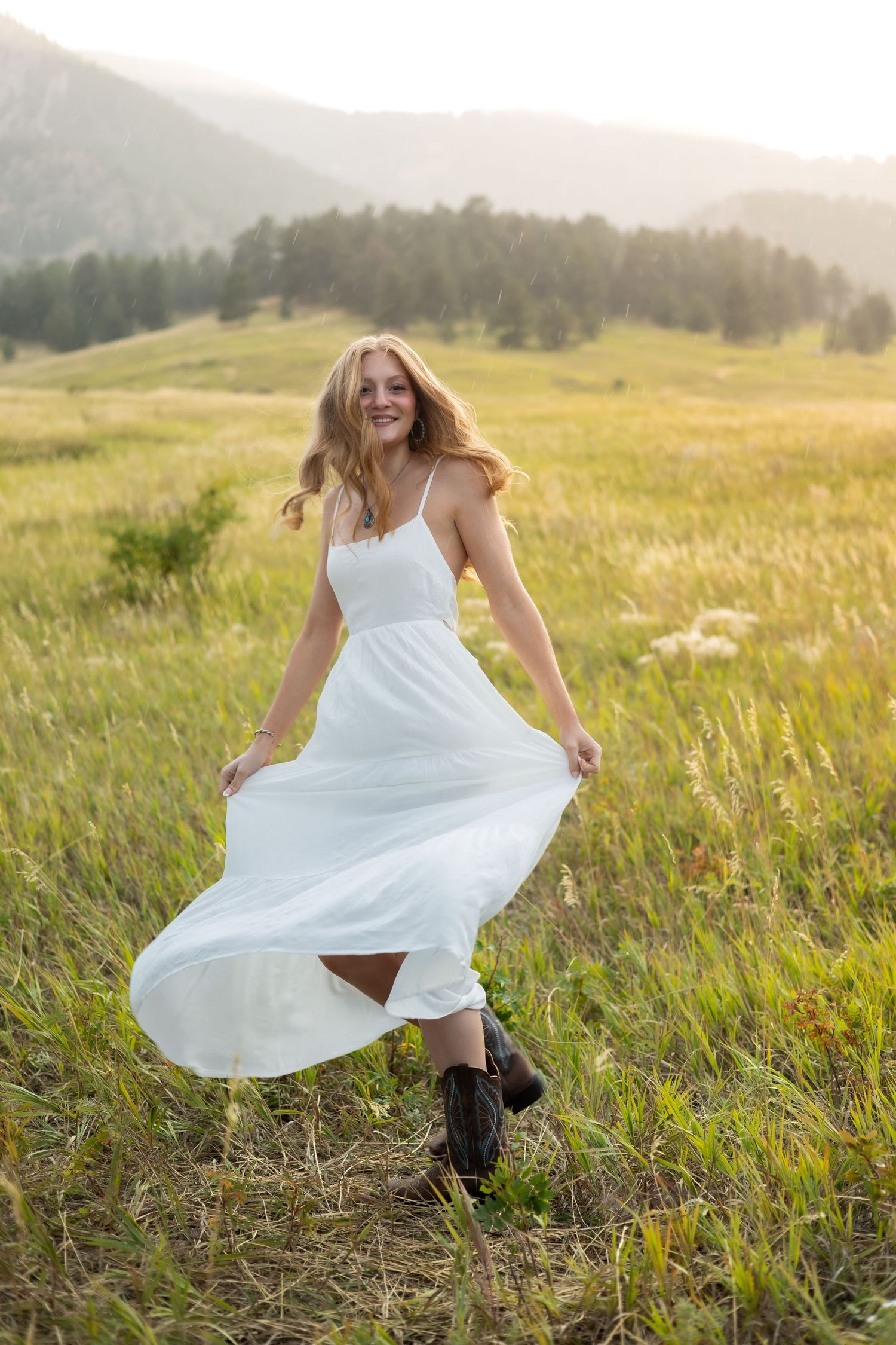 A white sundress twirls against a backdrop of mountains and tall grass at sunset.
