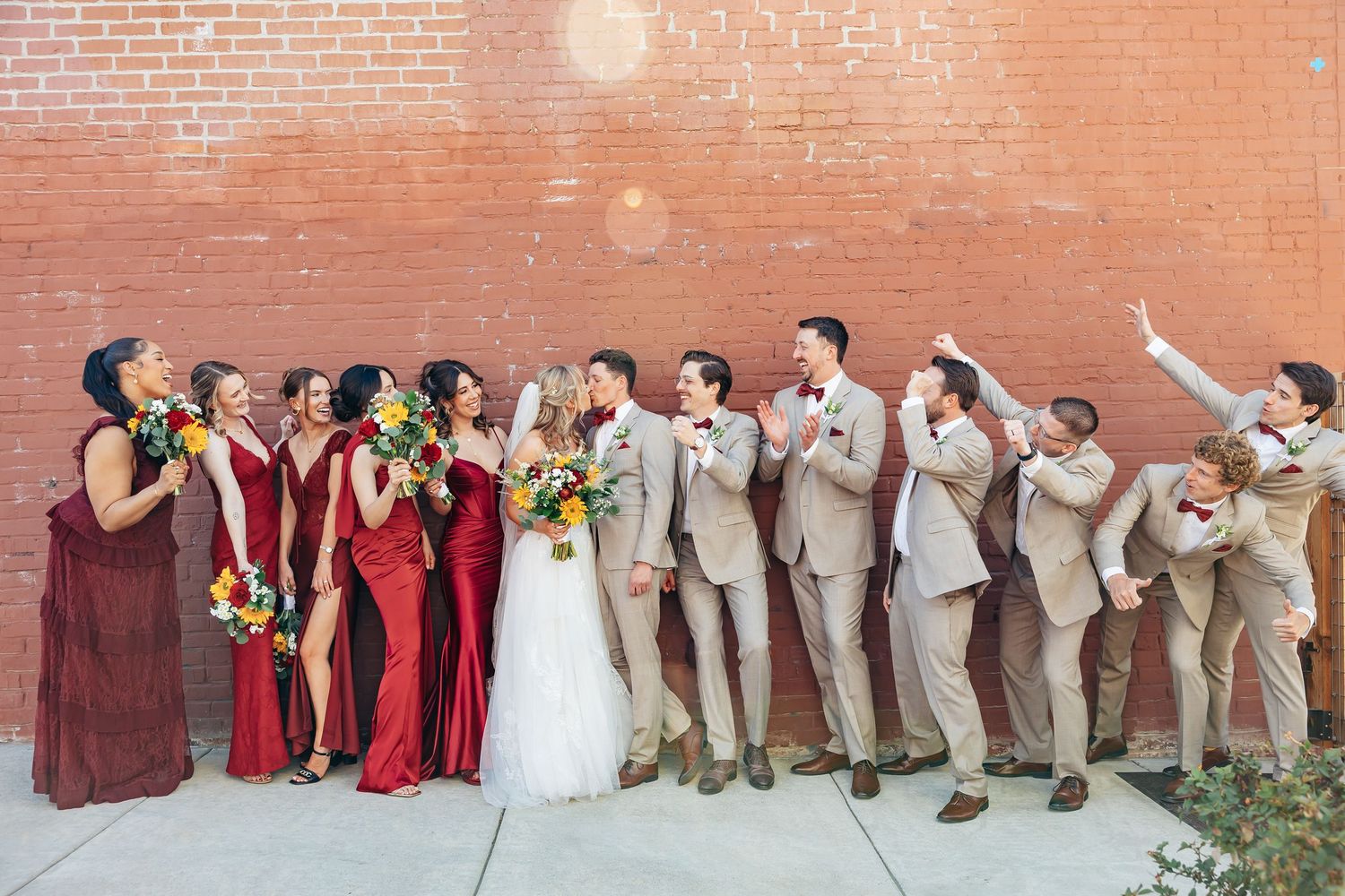 Wedding party in burgundy and tan suits poses laughing by brick wall with colorful bouquets.