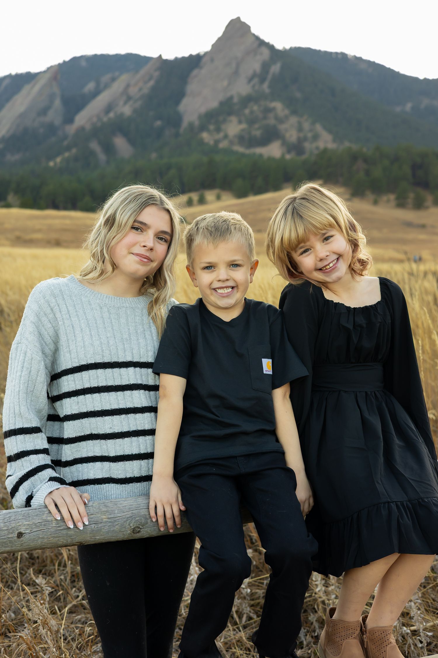 Three children sit together on a wooden fence with the Flatirons rock formation visible in Boulder, Colorado.