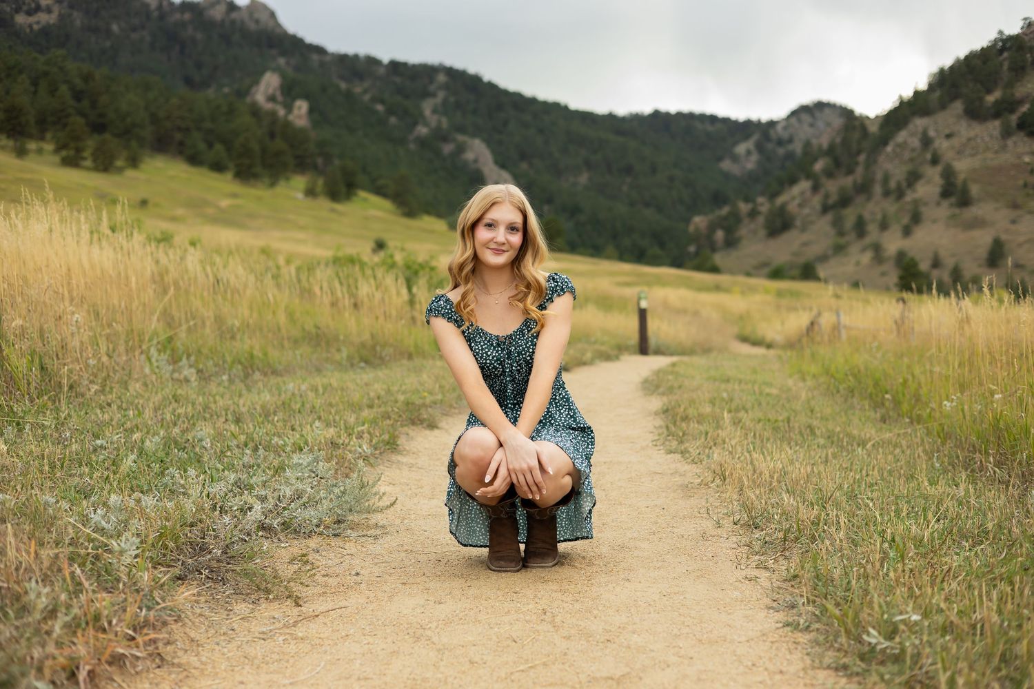 Young woman in teal dress crouching on dirt path surrounded by green meadows and rocky mountains.