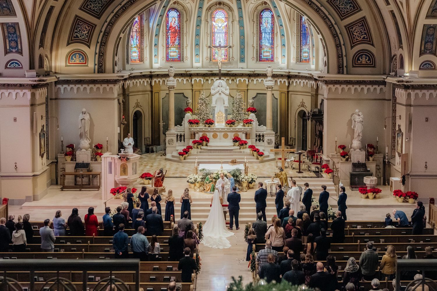 Elegant Catholic church wedding ceremony with bride and groom at altar, red floral decor, stained glass windows, and guests in pews.