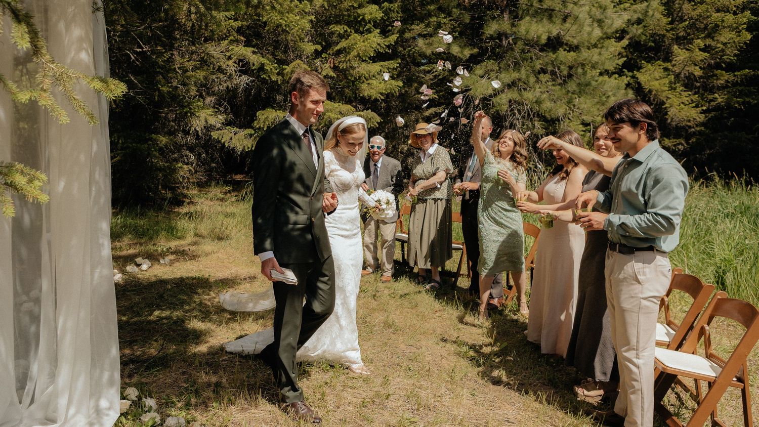 Wedding ceremony taking place in a wooded outdoor setting with guests seated on wooden benches.
